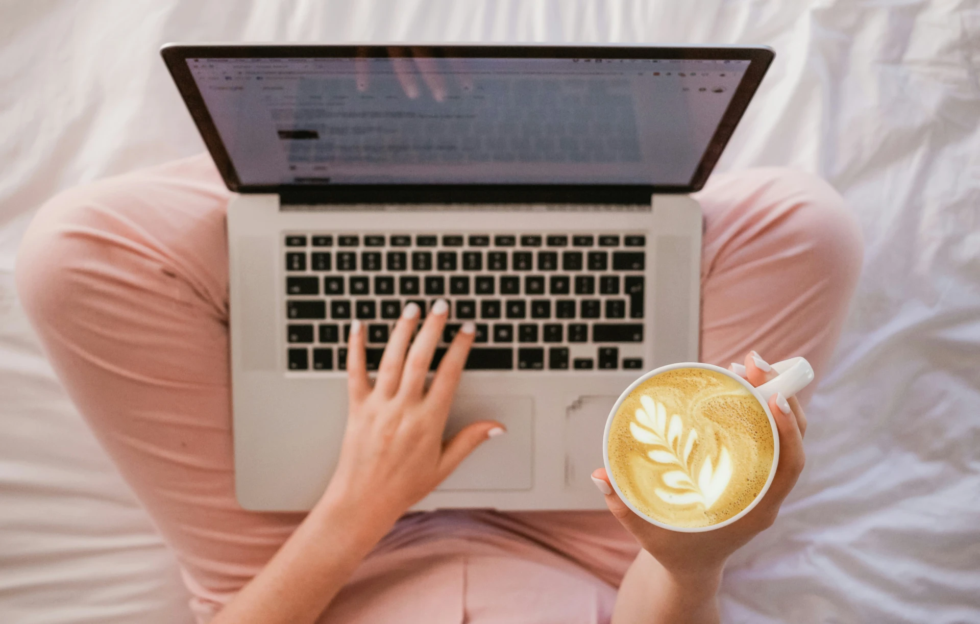 Person wearing pink pajamas types on a laptop while holding a cup of coffee on a white bed.