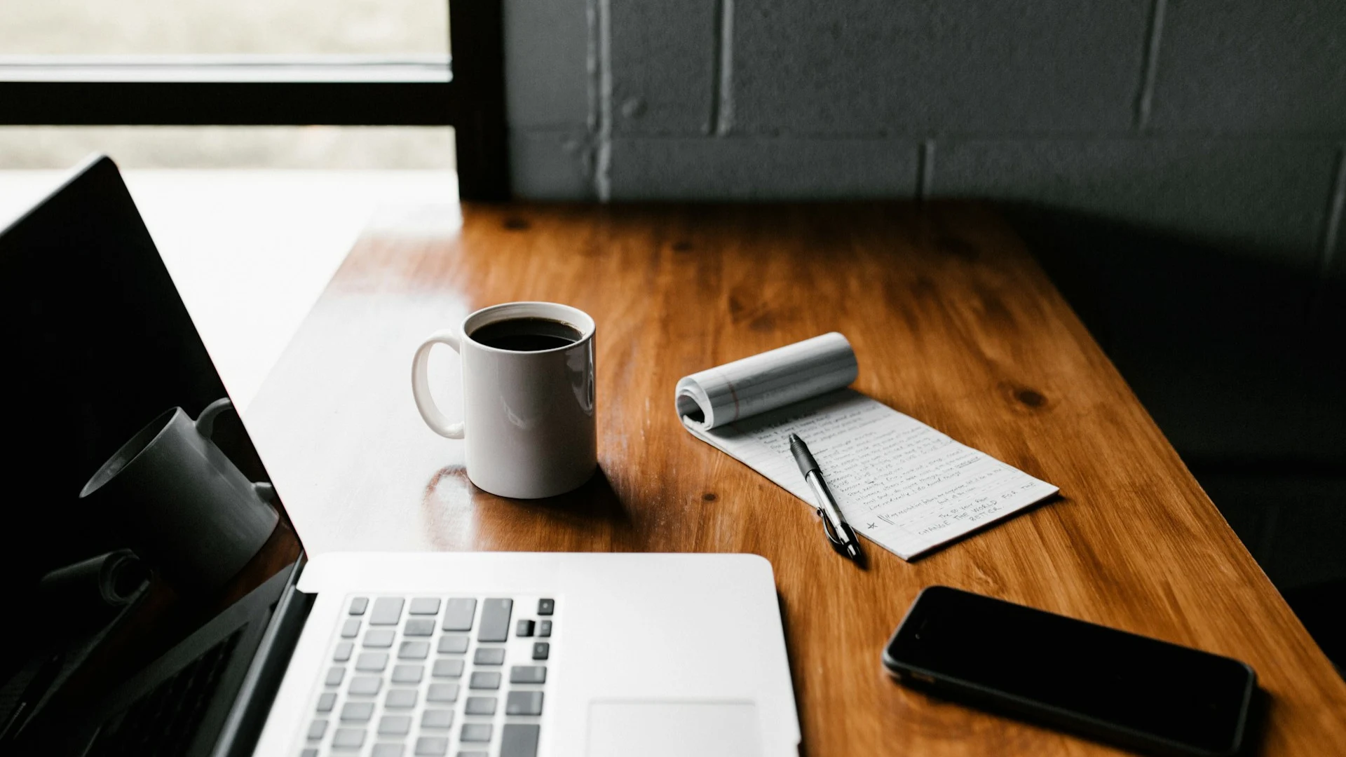 Laptop, coffee mug, notepad, pen, and smartphone on a wooden desk.