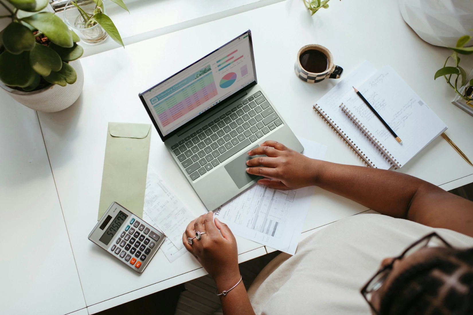 Overhead view of a person working on a laptop displaying financial data, surrounded by a calculator, notebook, coffee, and papers on a white desk.