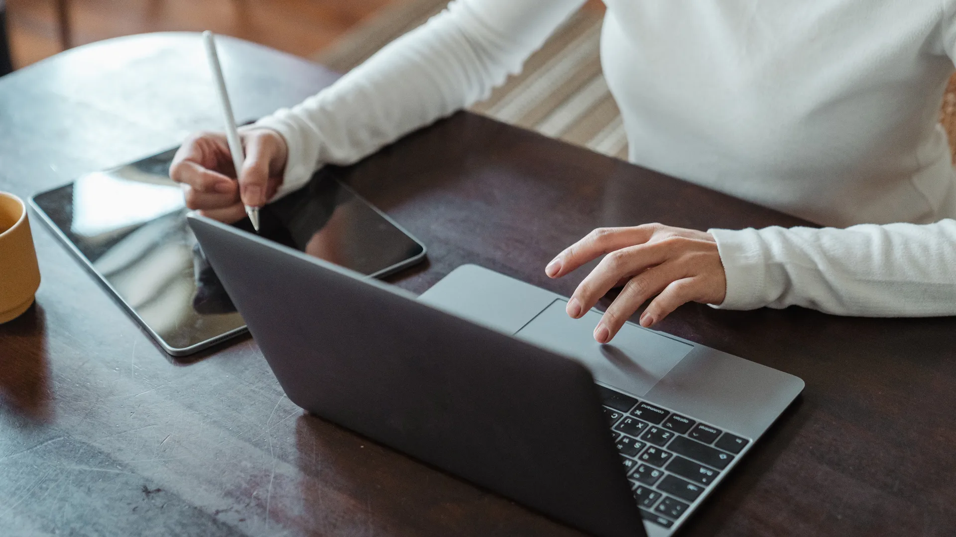 Person wearing white sleeves working on a laptop while simultaneously drawing on a tablet with a stylus at a dark wooden desk.