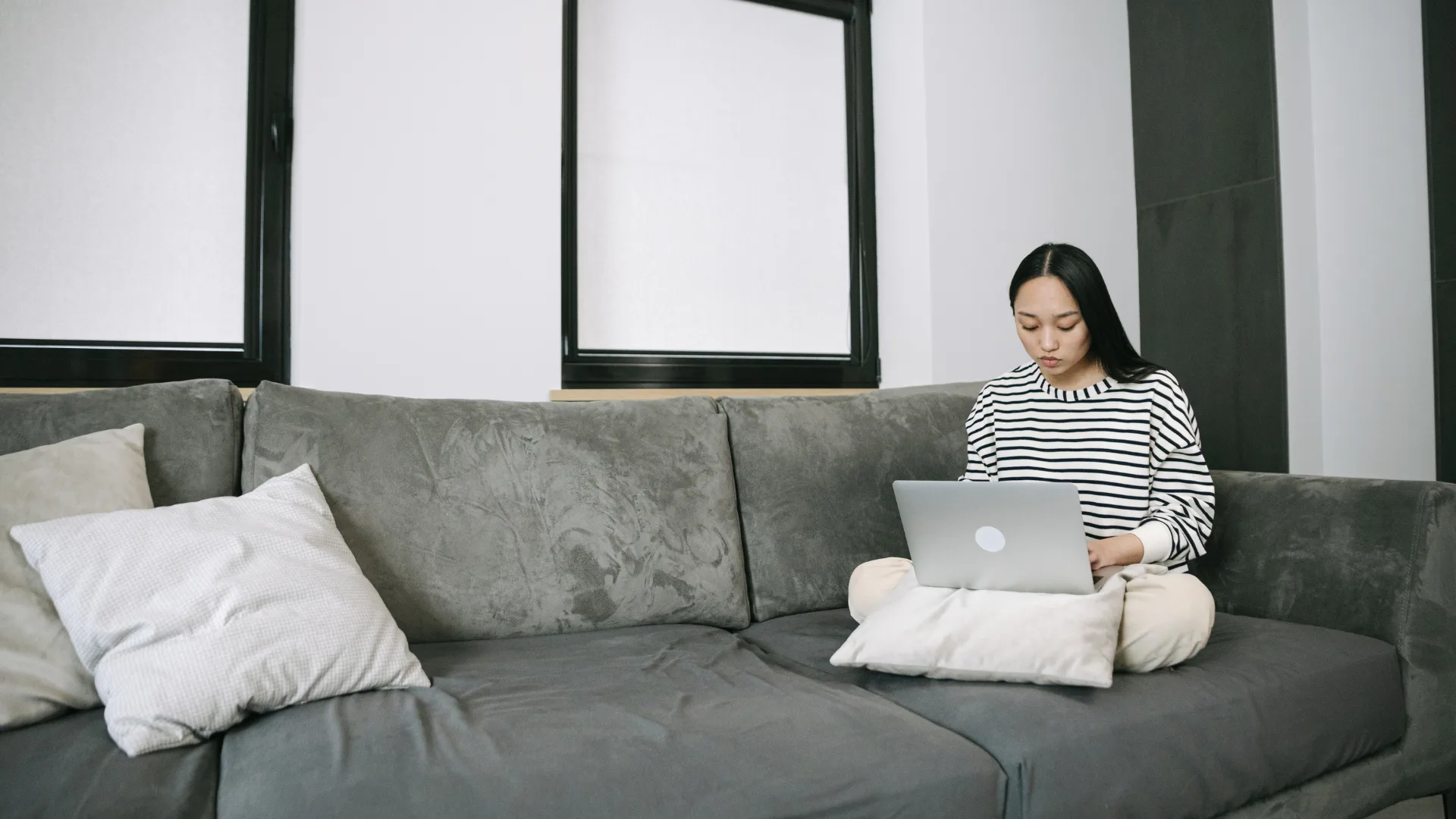 Young Asian woman with long dark hair wearing a striped shirt works on a silver laptop while sitting cross-legged on a dark gray sofa with white pillows.