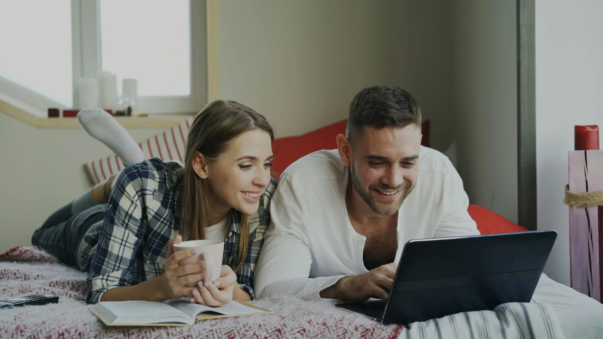A smiling couple lies on a bed looking at a laptop, with a book and mug nearby.
