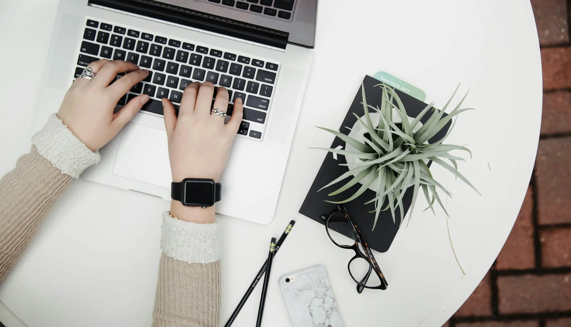 Hands typing on a laptop next to a potted plant, glasses, phone, and pencils on a white table.