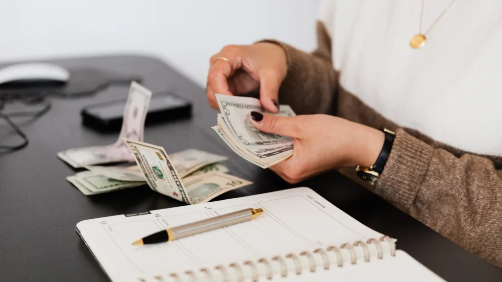 Person counting a fan of US dollar bills over an open planner with a pen resting on the page.