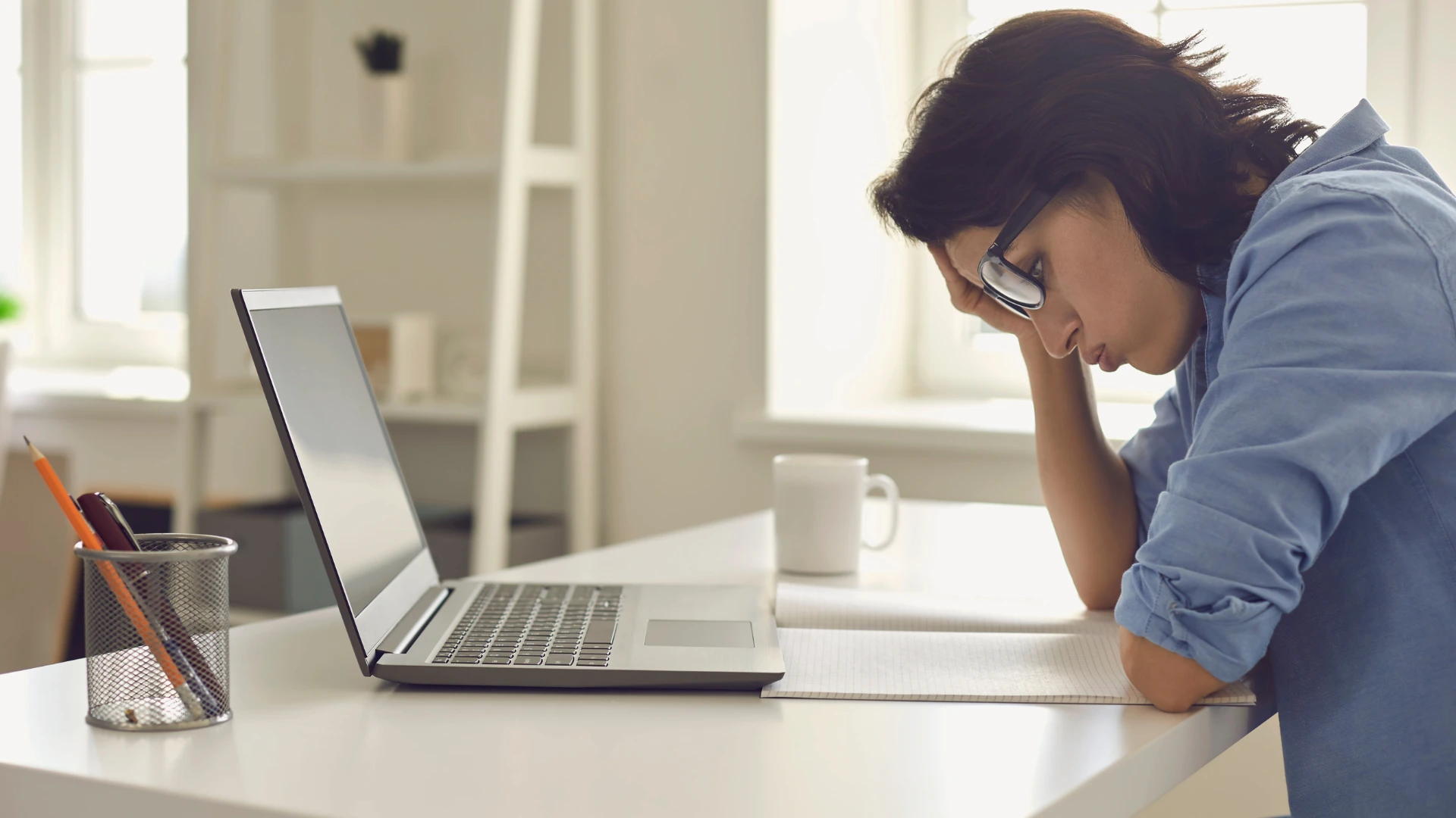 Woman wearing glasses leaning her head on her hand over an open notebook next to a laptop on a white desk.