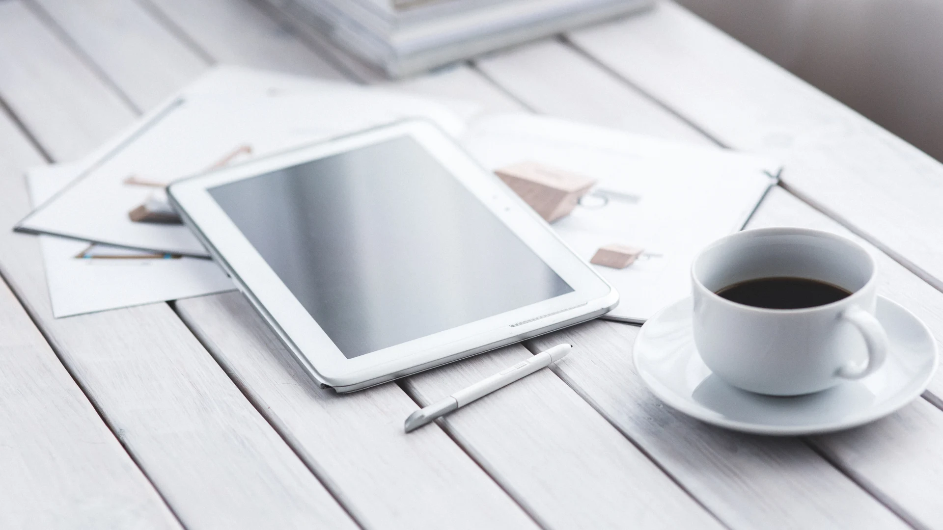 White tablet and a cup of black coffee resting on a light-colored wooden plank surface with scattered papers.