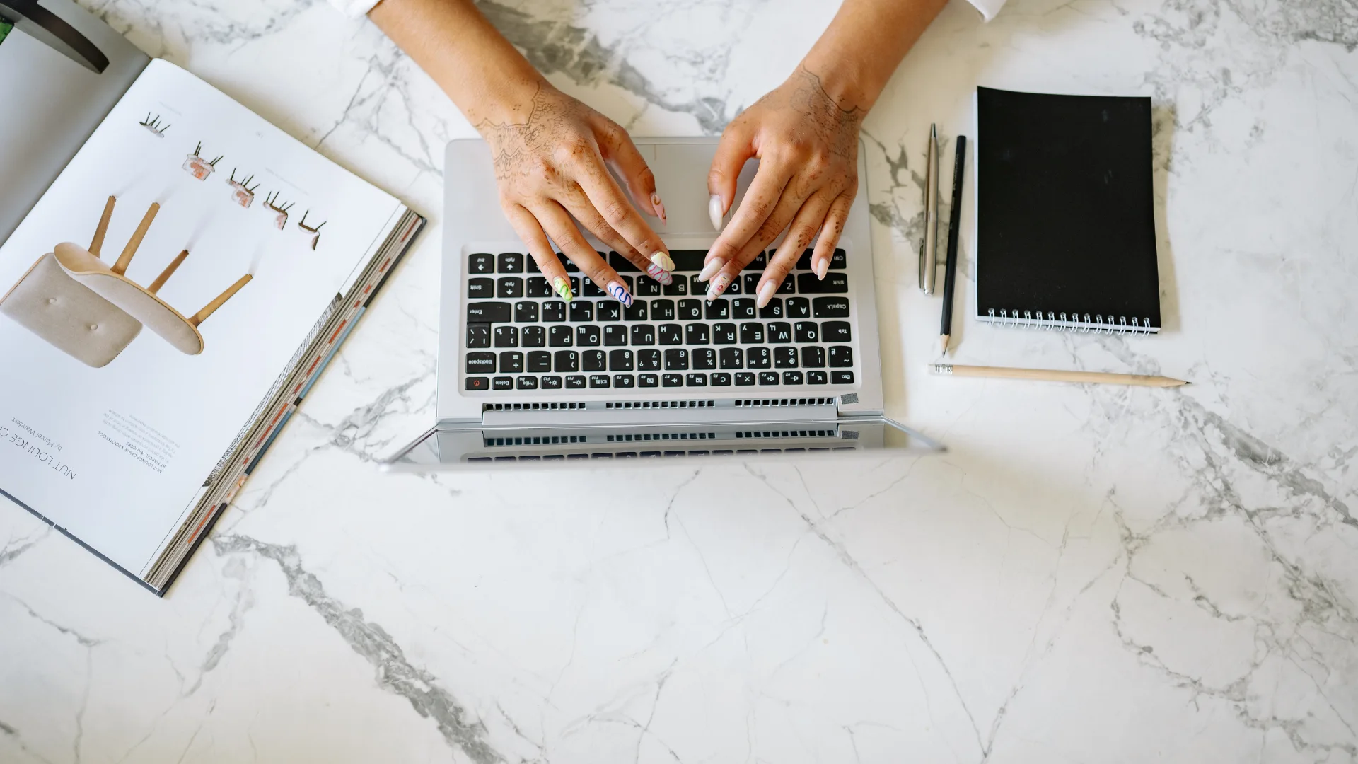 Overhead view of hands with decorative nails typing on a silver laptop next to an open design magazine and a black notebook on a white marble surface.