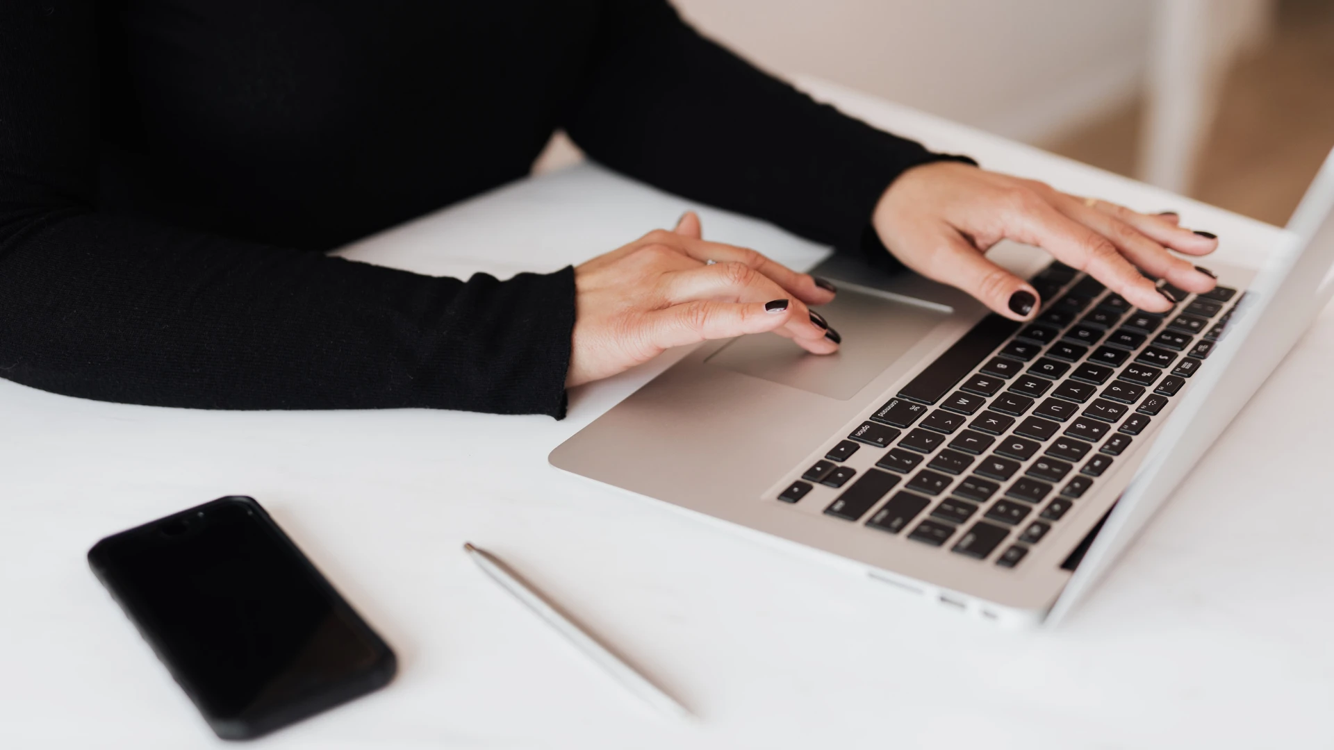 Close-up of a person wearing a black top using the trackpad and keyboard of a silver laptop on a white desk next to a black smartphone and a pen.