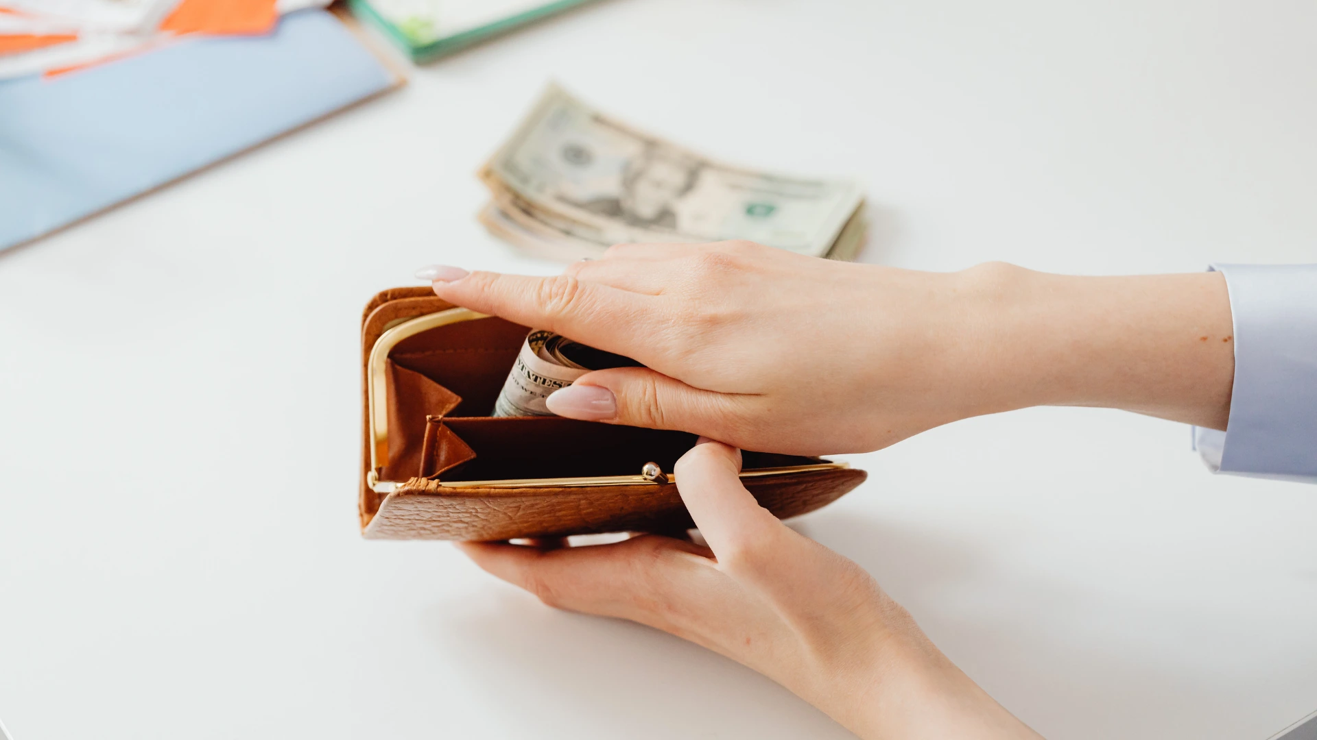 Hands place a rolled-up wad of US currency into a brown leather coin purse on a white desk with scattered twenty-dollar bills in the background.