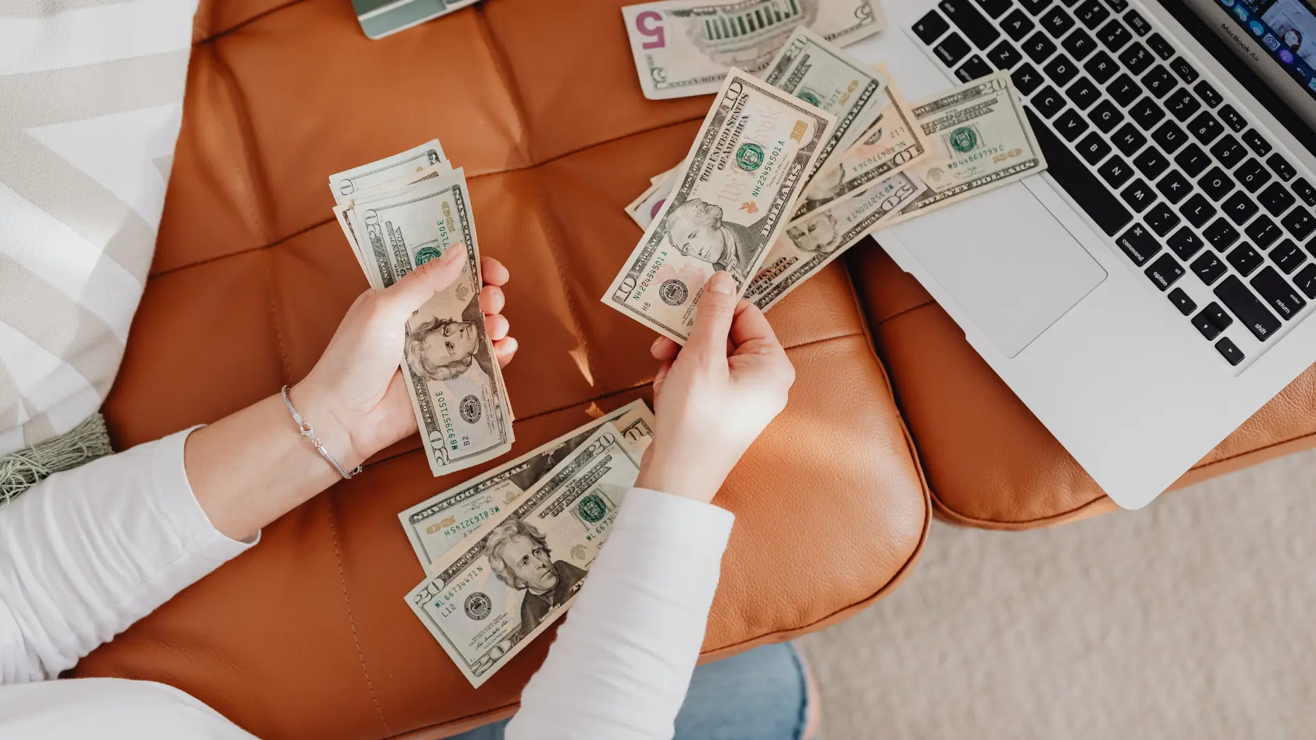 Person counting various denominations of US currency next to a silver laptop on a brown leather surface.