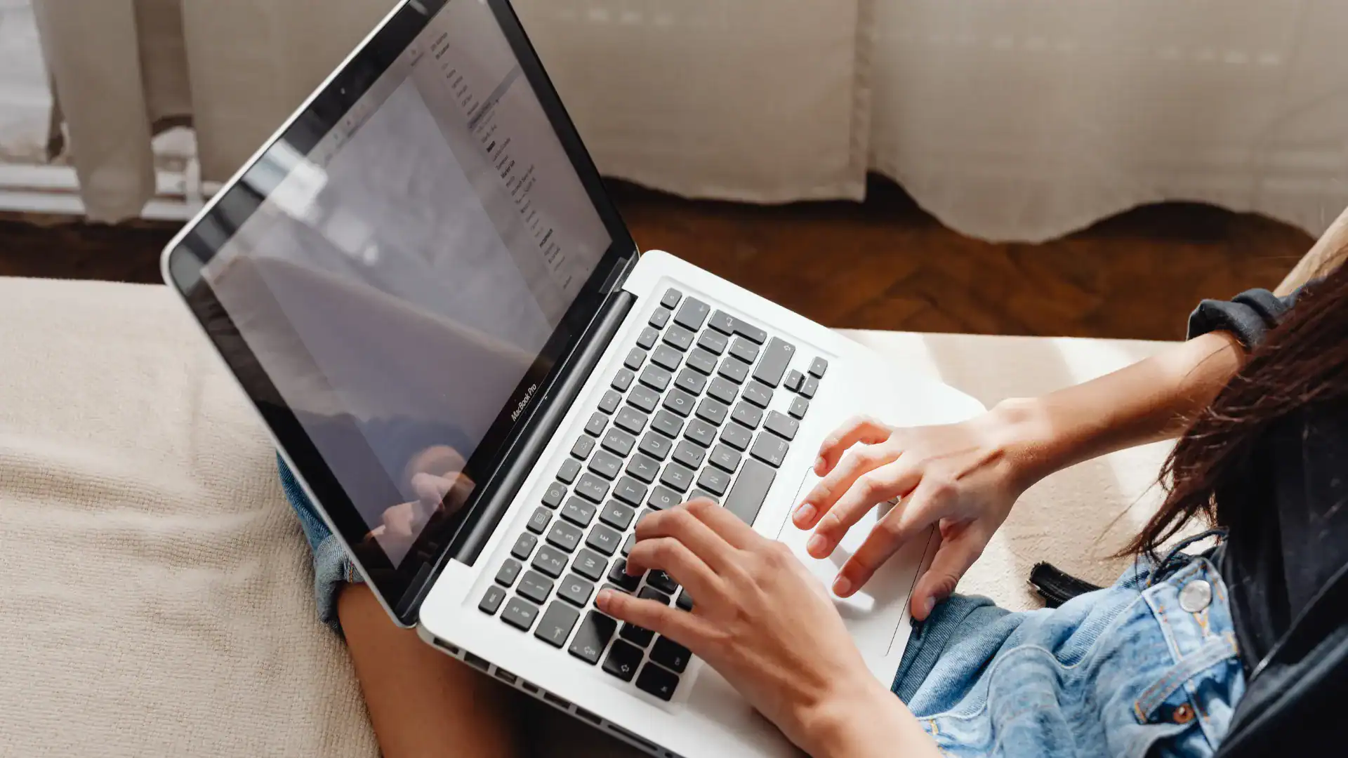 Person typing on a silver laptop resting on their lap while sitting indoors on a light-colored textured surface.