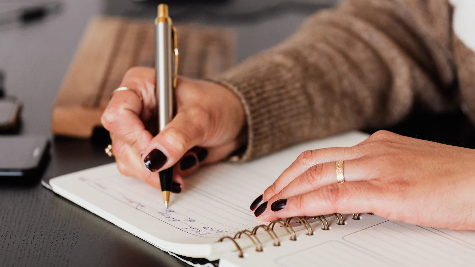 Close-up of a person wearing a brown sweater writing in a spiral-bound notebook with a silver and gold pen.