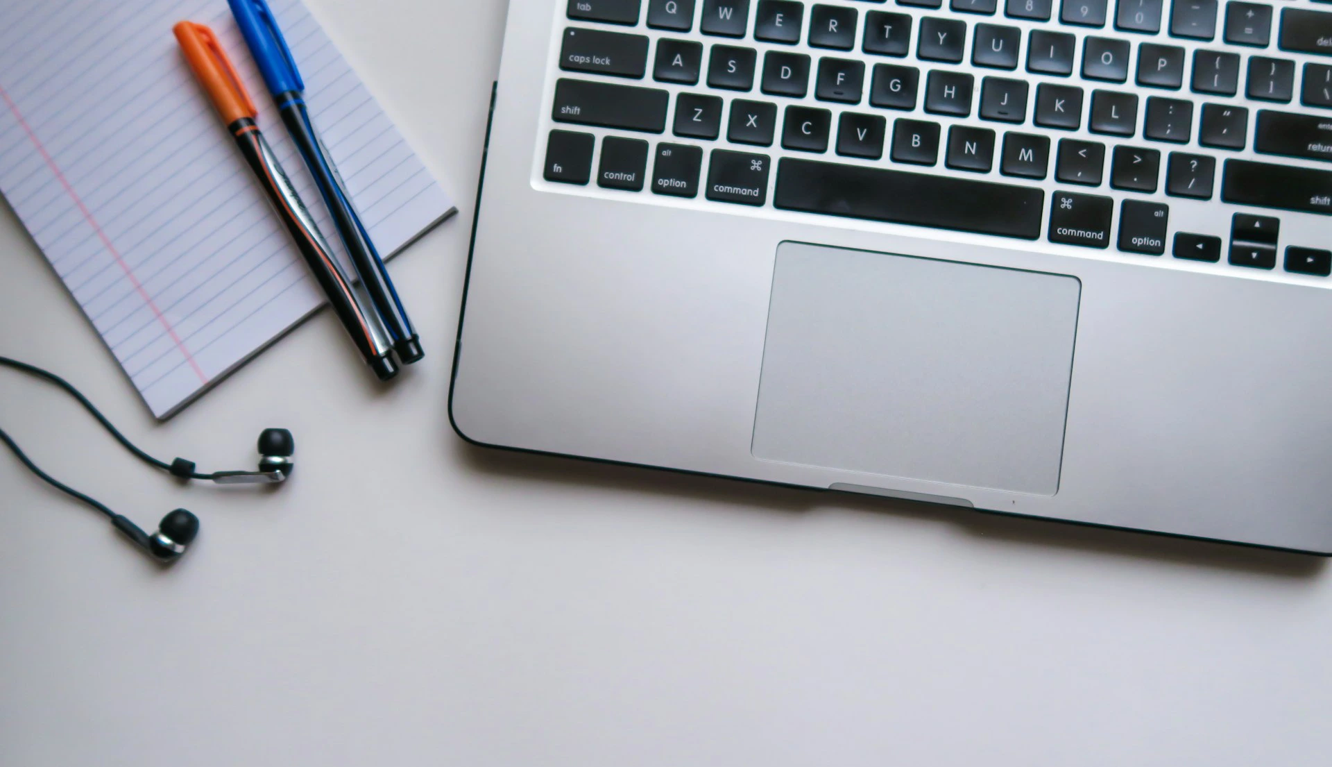 Laptop, notebook, pens, and earbuds arranged on a white surface.