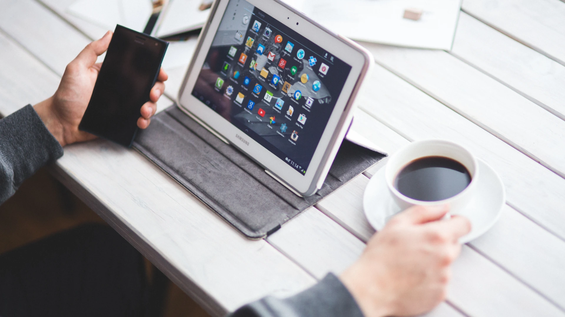 Person holding a smartphone and a cup of coffee next to a tablet displaying app icons on a white wooden slatted table.