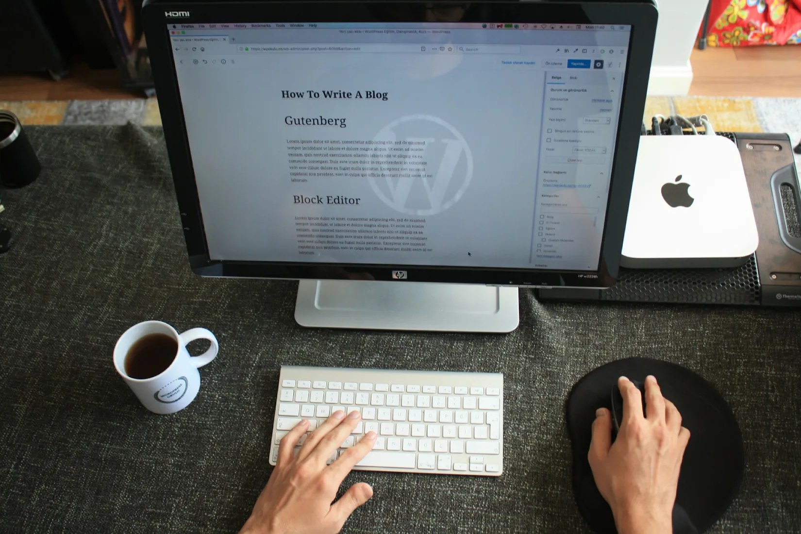 Overhead view of hands typing on a white keyboard while editing a WordPress blog post titled "How To Write A Blog" on an external monitor next to an Apple Mac Mini and a cup of coffee.