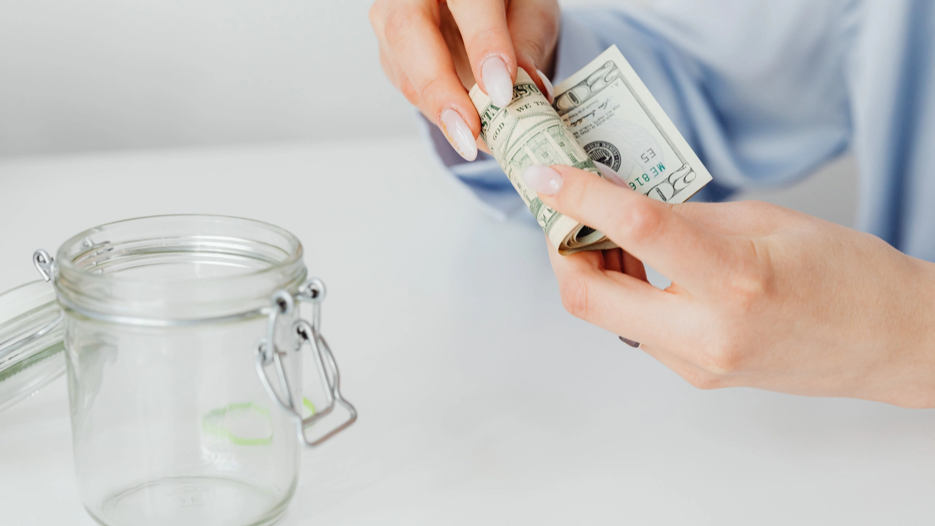 Woman's hands roll a U.S. twenty-dollar bill next to an empty glass canning jar on a white surface.