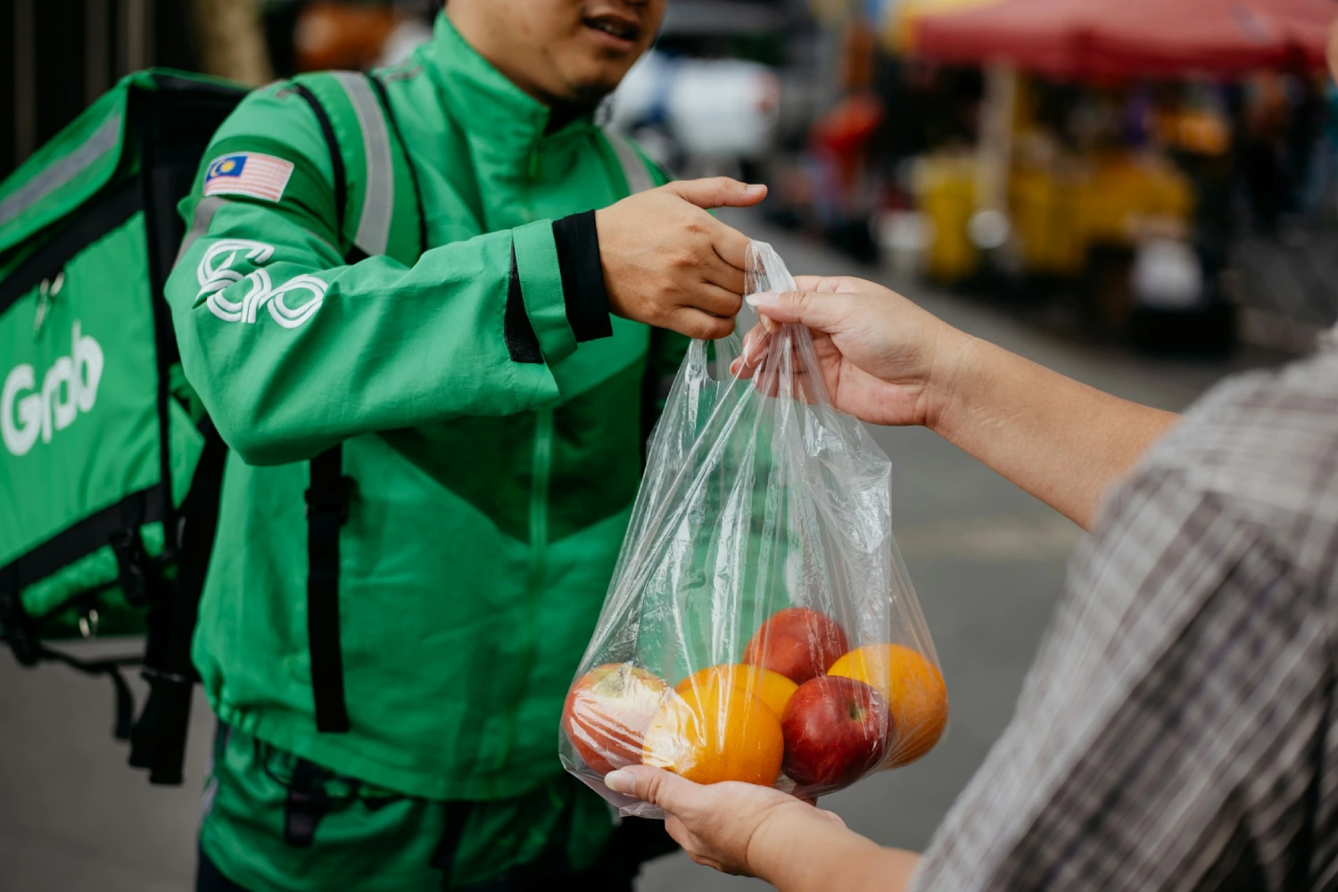 A Grab delivery driver in a green uniform hands a customer a plastic bag containing apples and oranges outdoors.