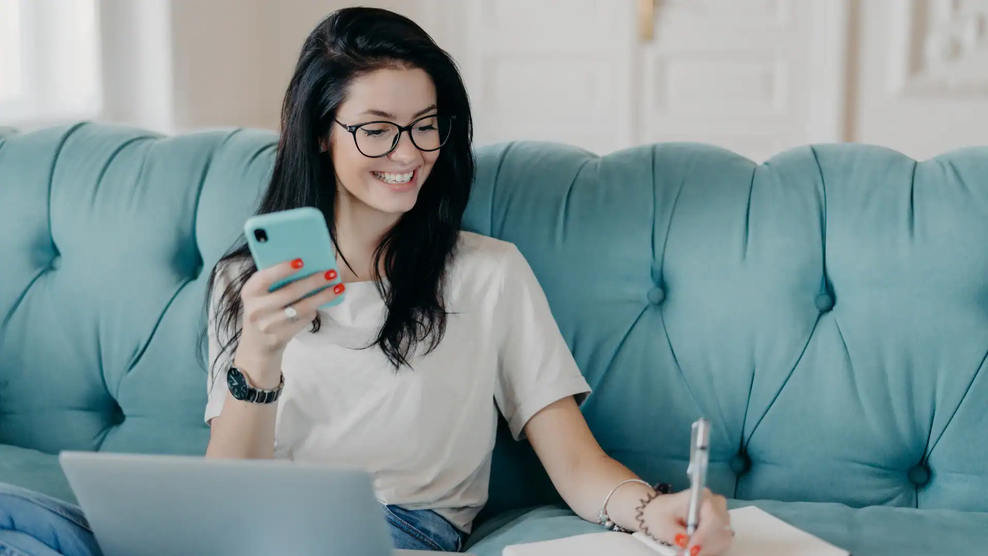 Smiling young woman with glasses looks at her phone while sitting on a teal tufted sofa next to a laptop and taking notes in a book.