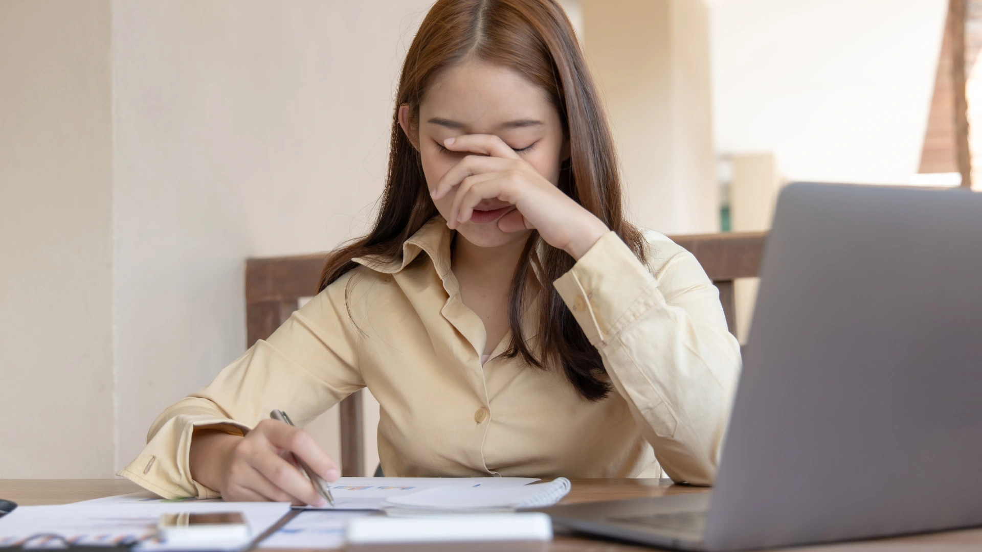 Tired young Asian woman in a beige shirt rubs her eyes while sitting at a desk with papers and a laptop.