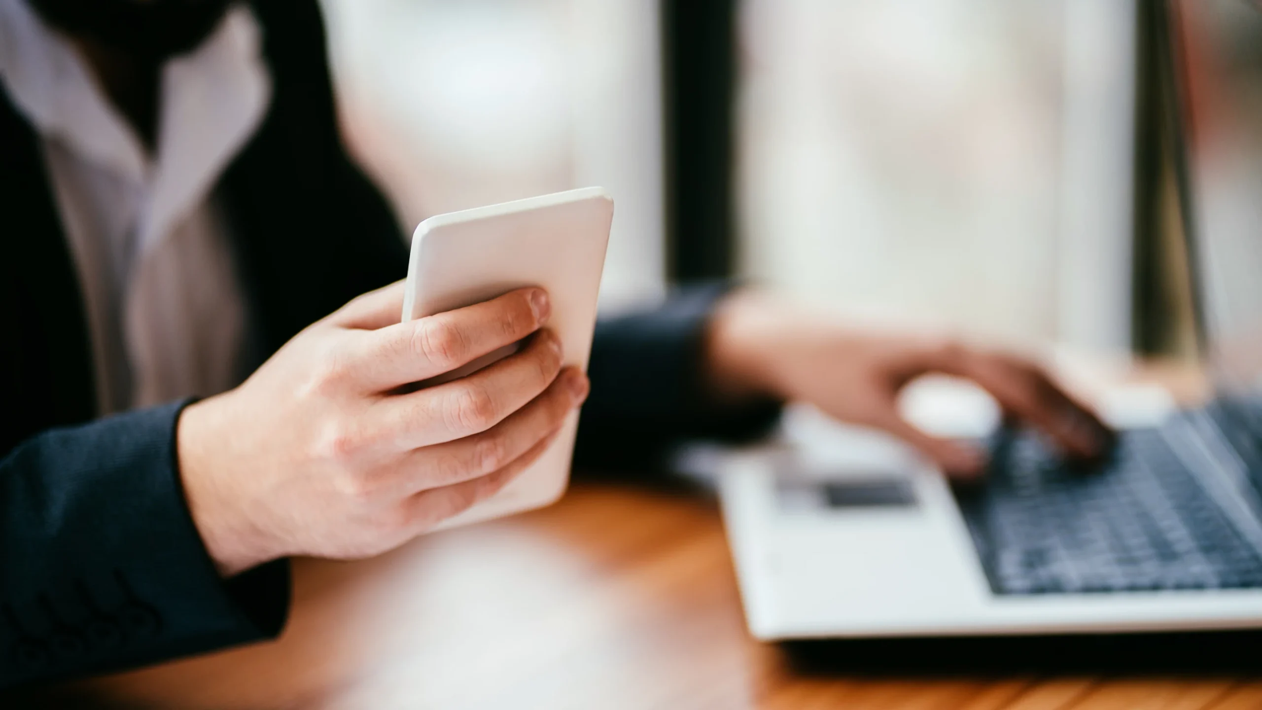 Close-up of a person in business attire holding a white smartphone while simultaneously typing on an open laptop.