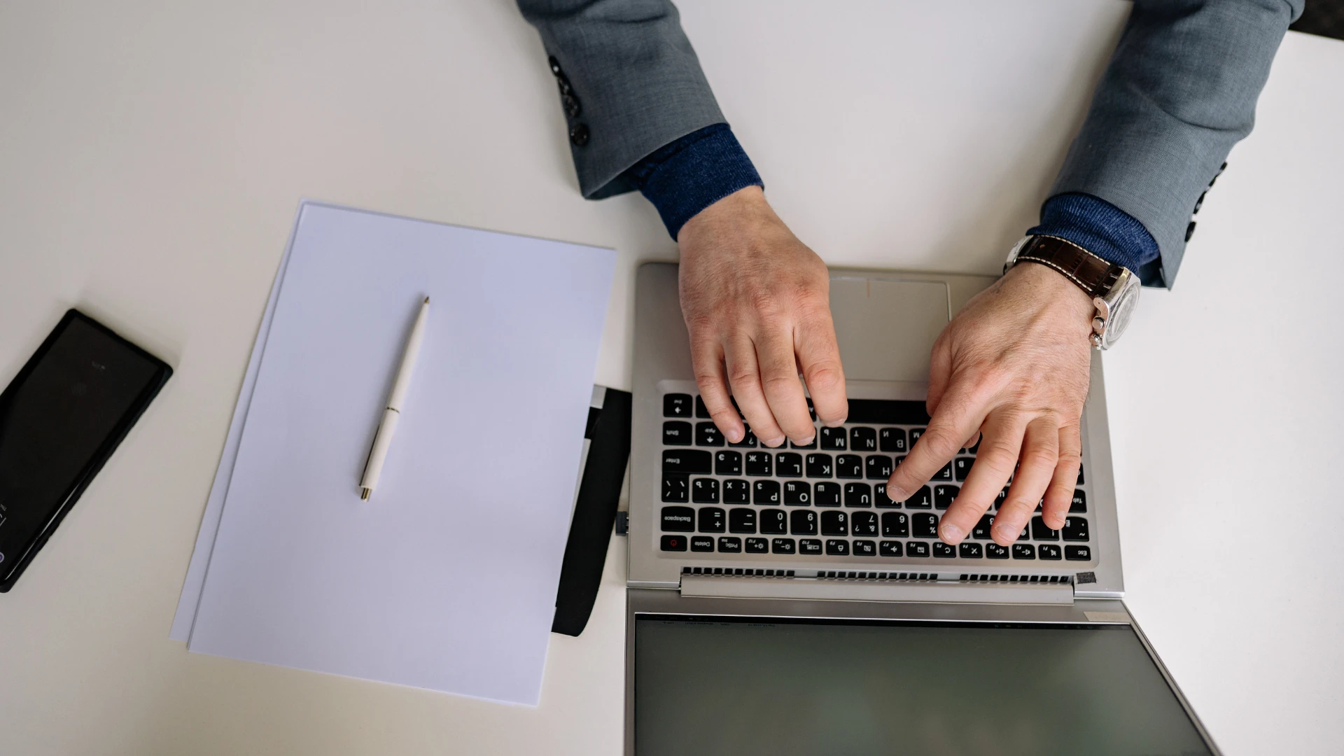 Overhead view of a person in a gray jacket typing on a silver laptop next to stacked papers, a white pen, and a black smartphone on a white desk.