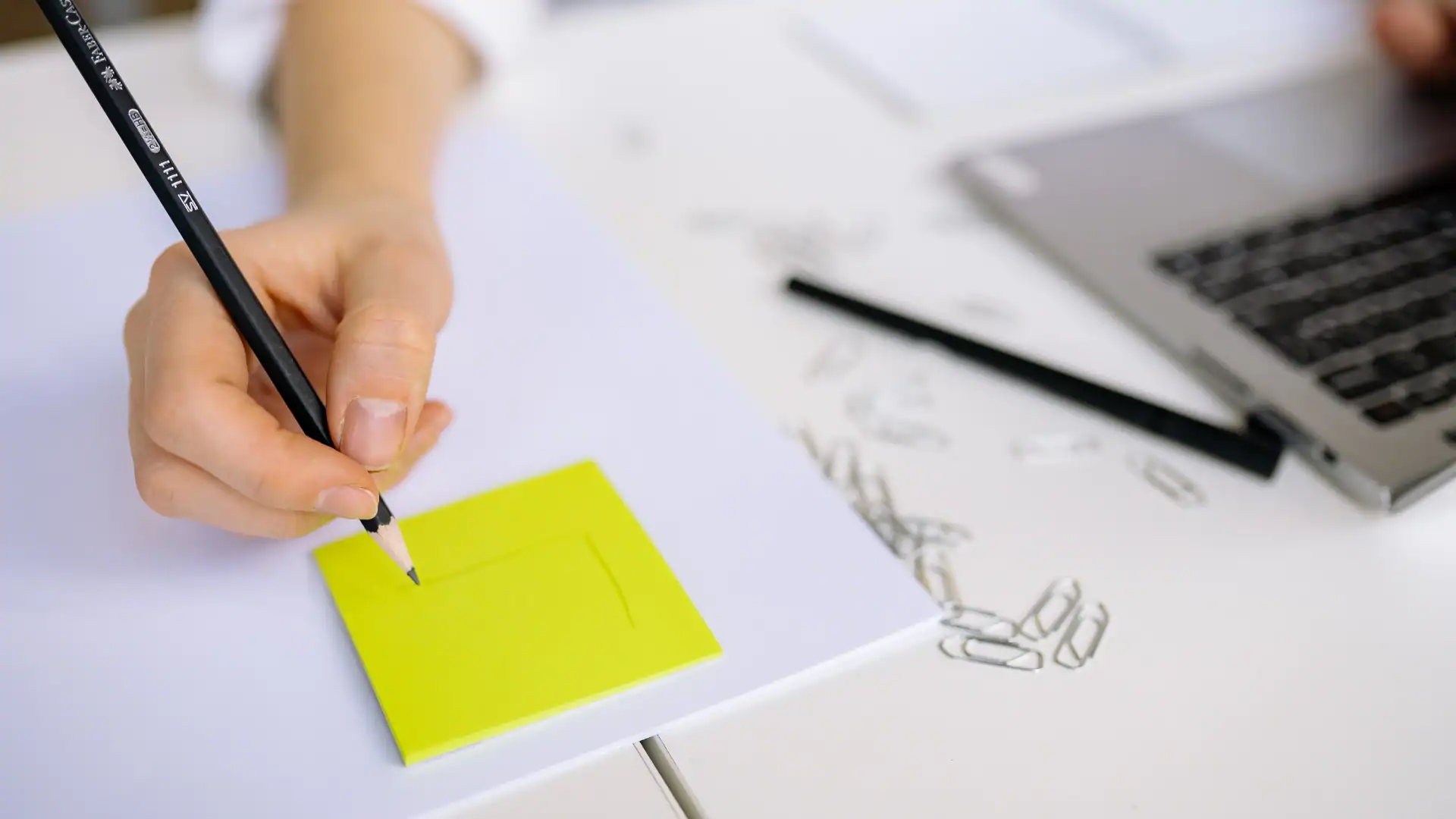 A person's hand holding a black pencil writing on a bright yellow sticky note placed over white paper next to a silver laptop.