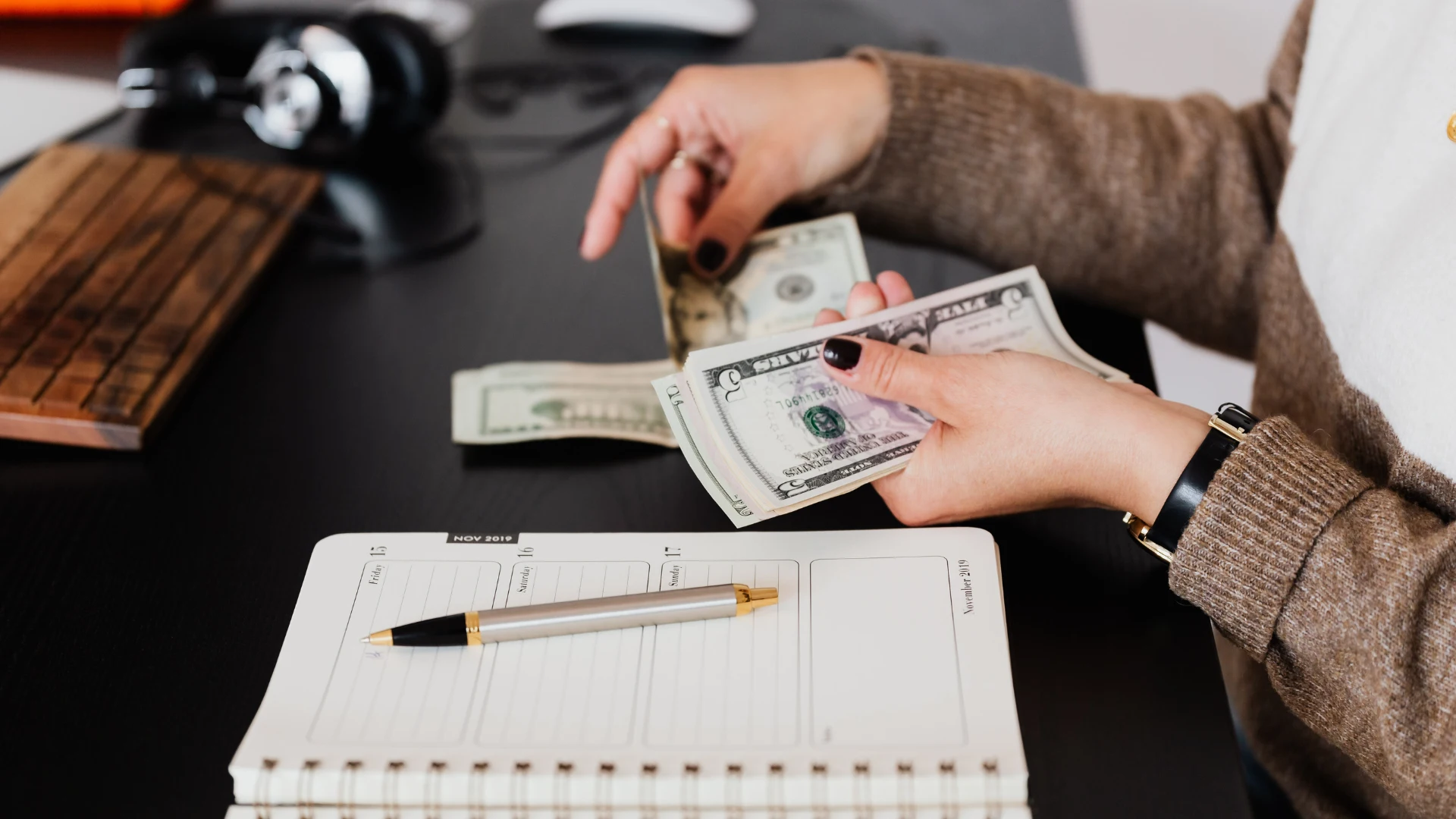 Woman in a brown sweater counting US dollar bills over an open planner with a silver pen resting on the page.