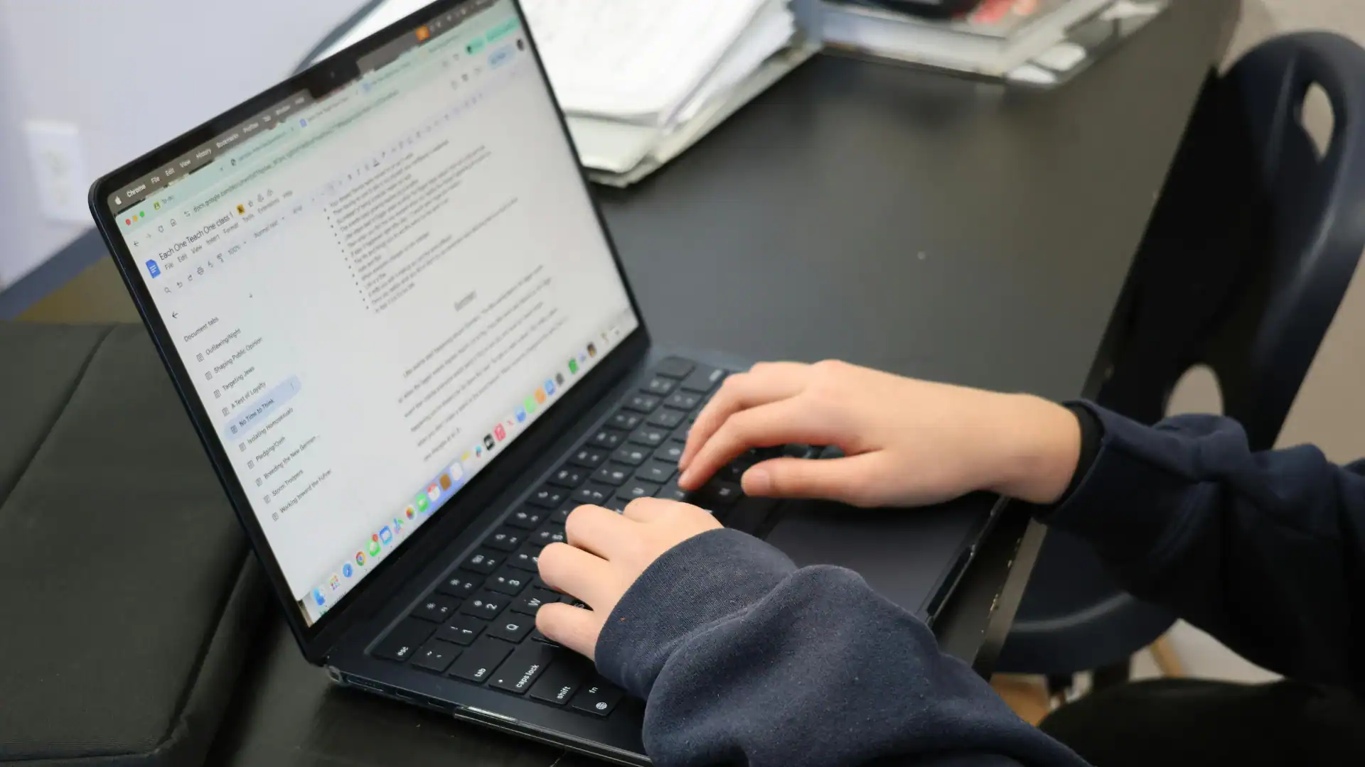 Close-up view of hands wearing a dark blue sweatshirt typing on a laptop displaying a document in Google Docs at a dark desk.