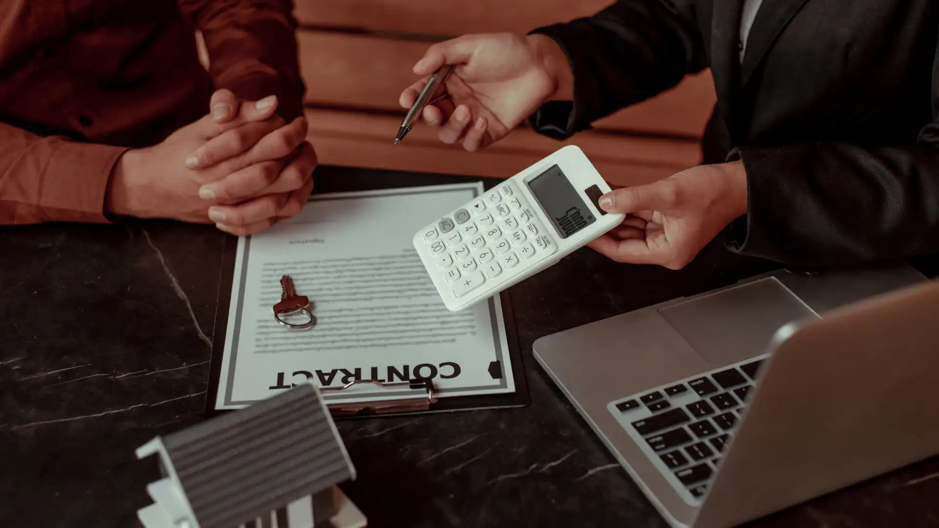 Two people review a contract with a house key, calculator, and laptop on a dark marble table, suggesting a real estate transaction.