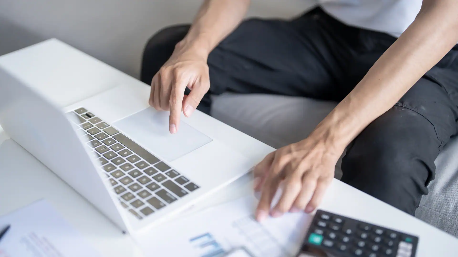 Close-up of a person using the touchpad on a white laptop while financial documents and a calculator rest on a white surface nearby.