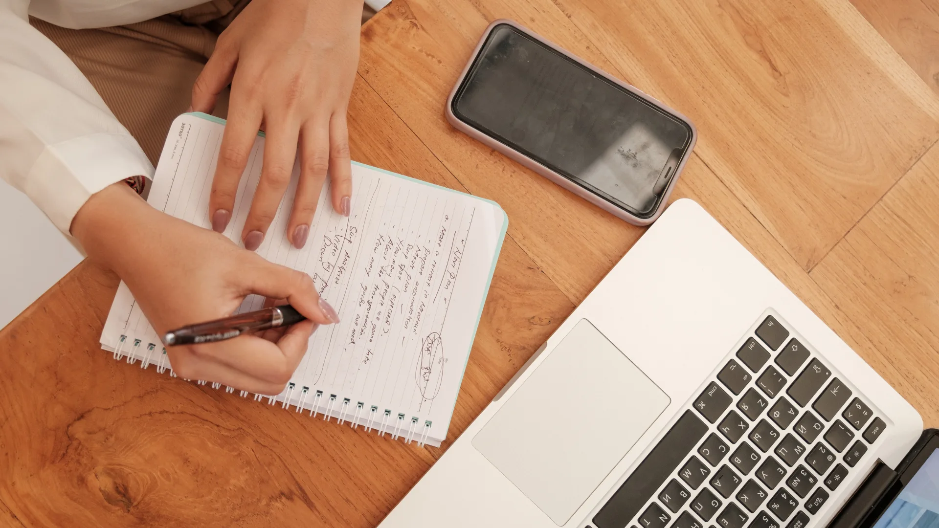 Overhead view of hands writing notes in a spiral notebook beside a laptop and smartphone on a wooden desk.