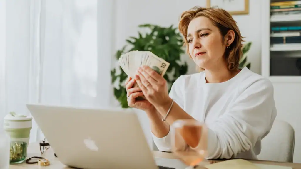 Woman in white shirt counting a fan of US dollar bills while sitting at a desk with a laptop open.
