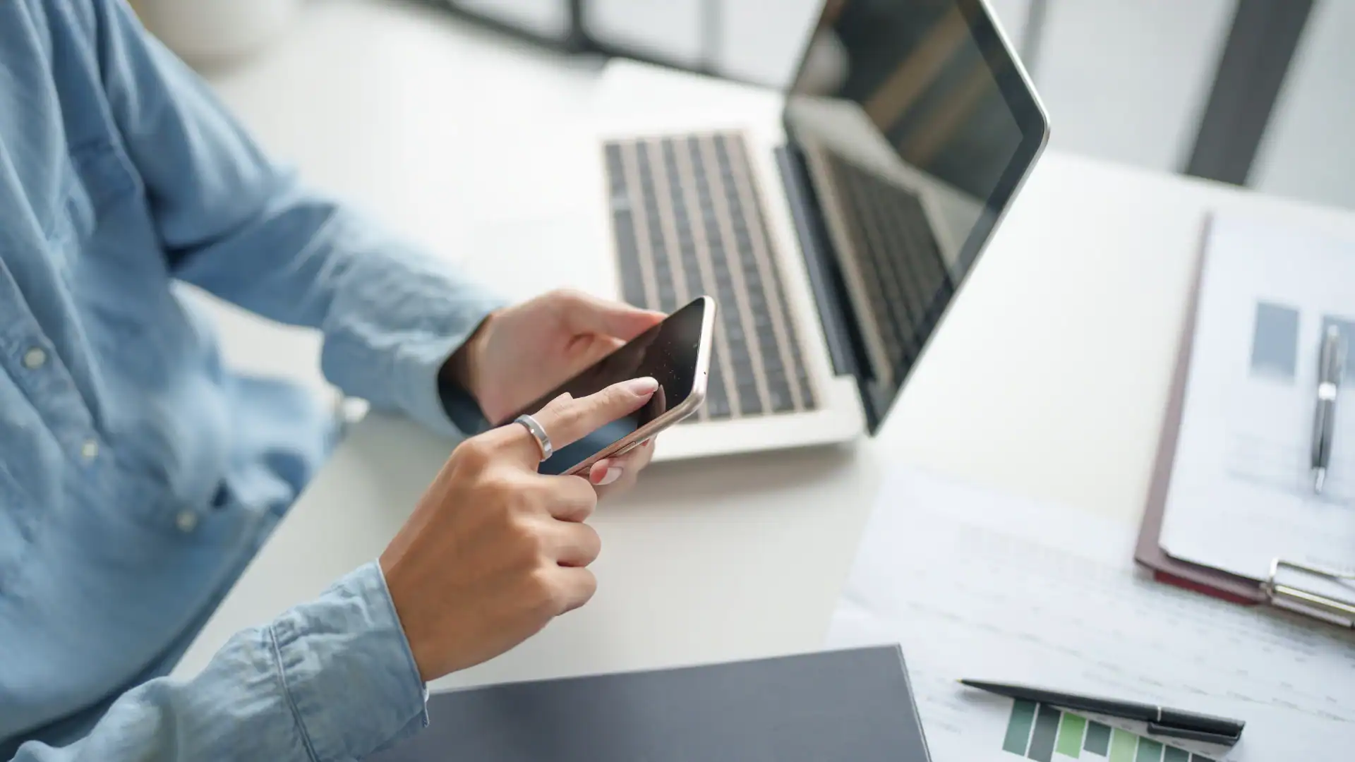 Person in a blue shirt using a smartphone at a white desk with a laptop and business documents visible.