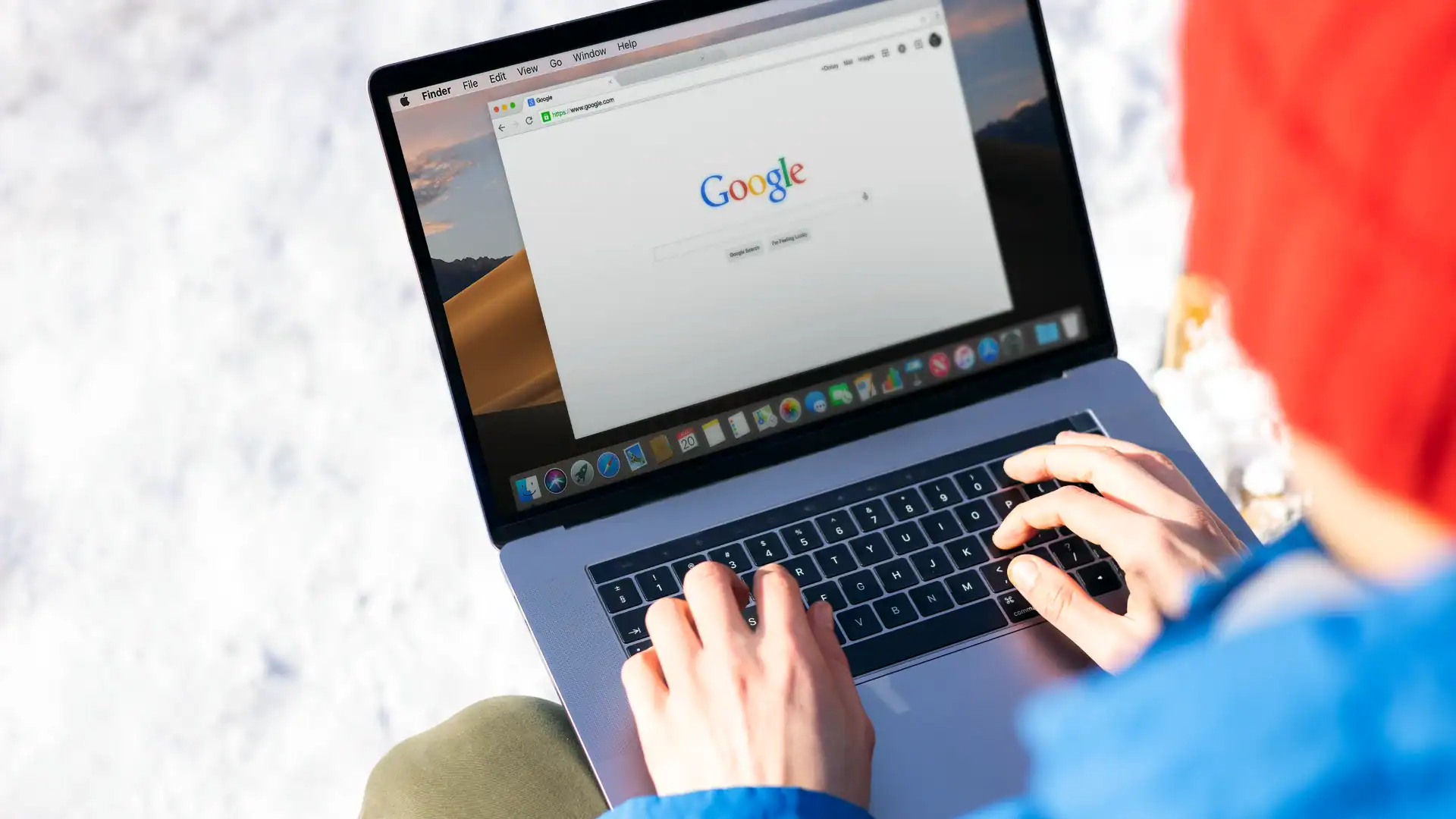 Person wearing a red hat uses a MacBook outdoors with a Google search page open on the screen.