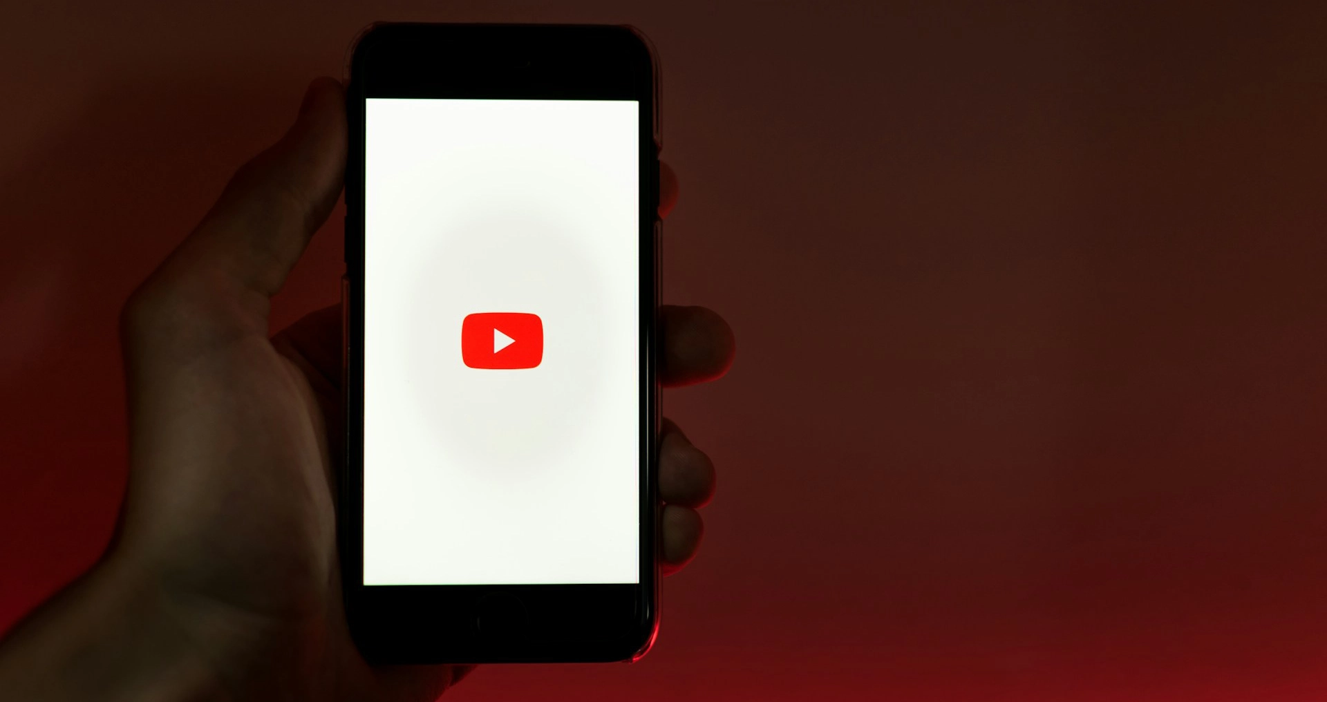 Hand holding a smartphone displaying the white YouTube loading screen with the red play button icon against a dark red background.