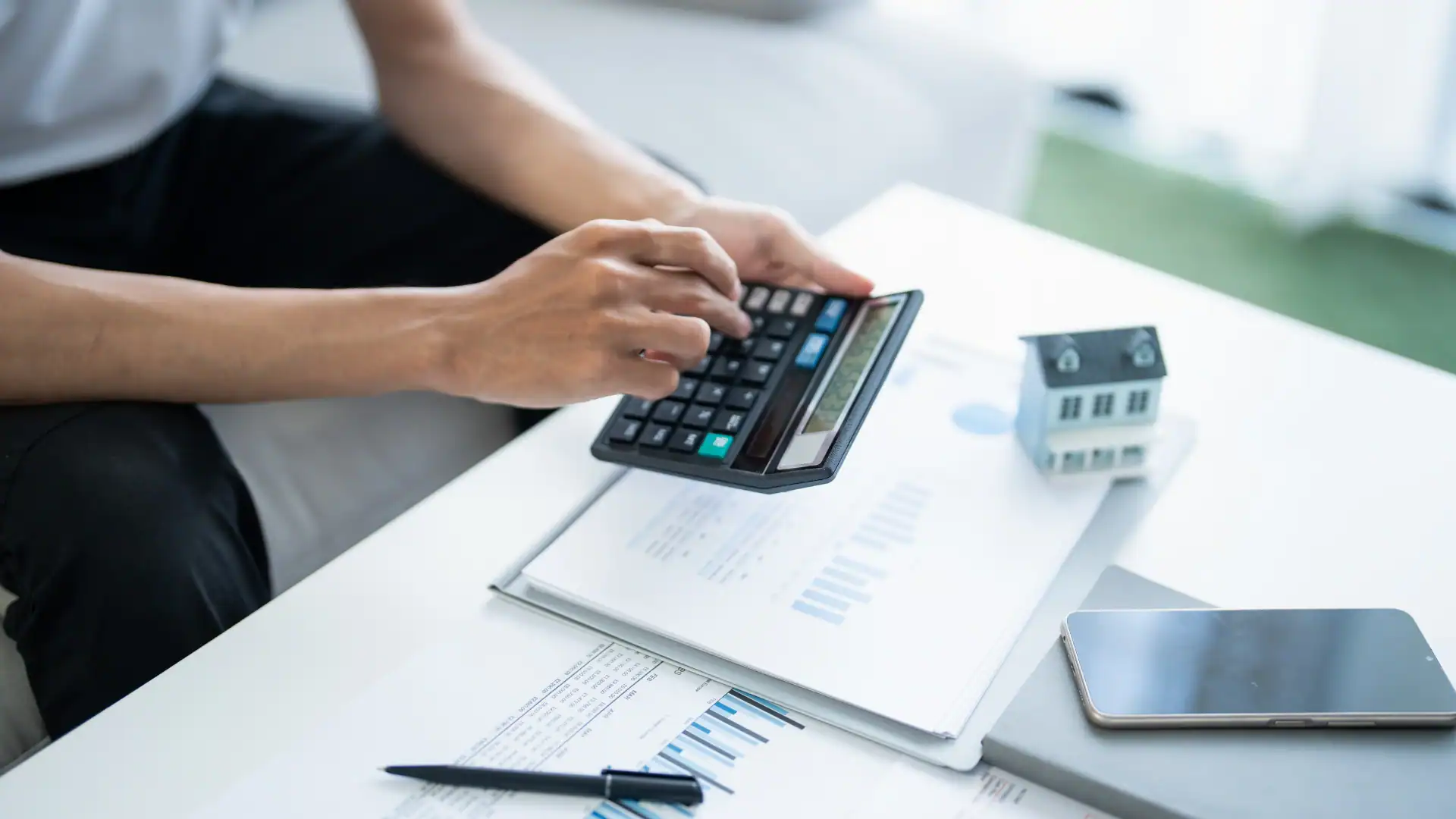 Person calculating finances using a calculator over documents with a miniature house model and smartphone nearby.