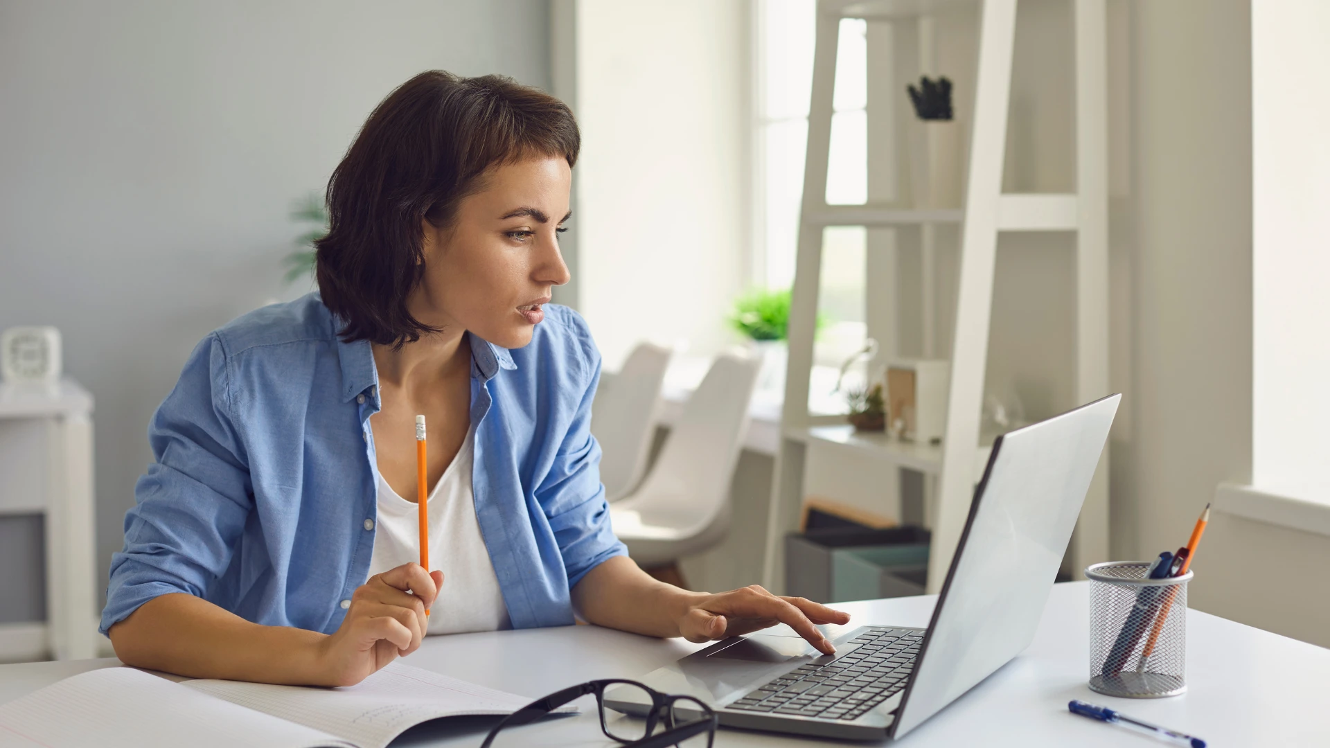 Young woman with short brown hair, wearing a blue button-down shirt, works on a laptop at a white desk.