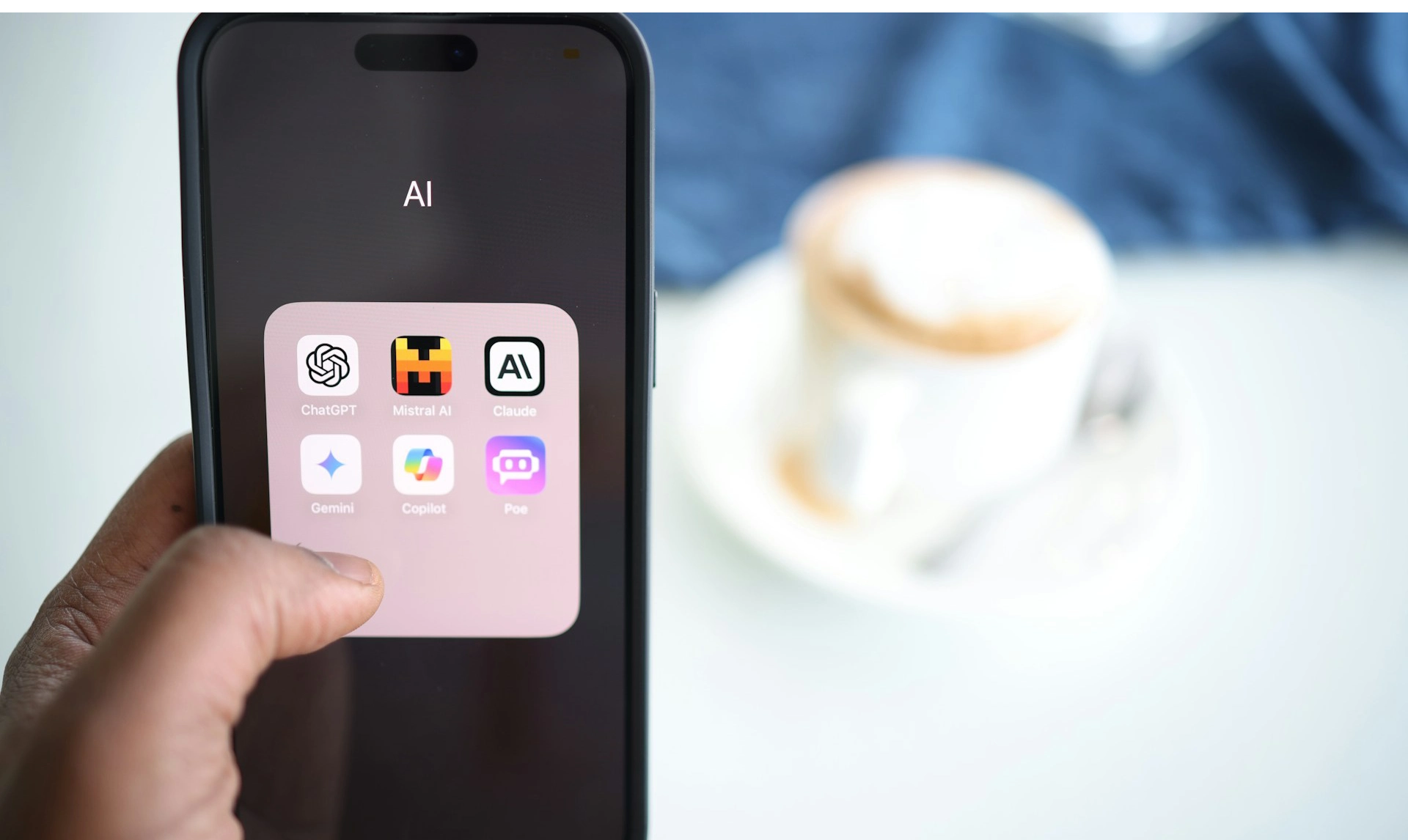 A hand holds a smartphone displaying the "AI" app folder with icons for ChatGPT, Mistral AI, Claude, Gemini, Copilot, and Poe, with a blurred coffee cup in the background.