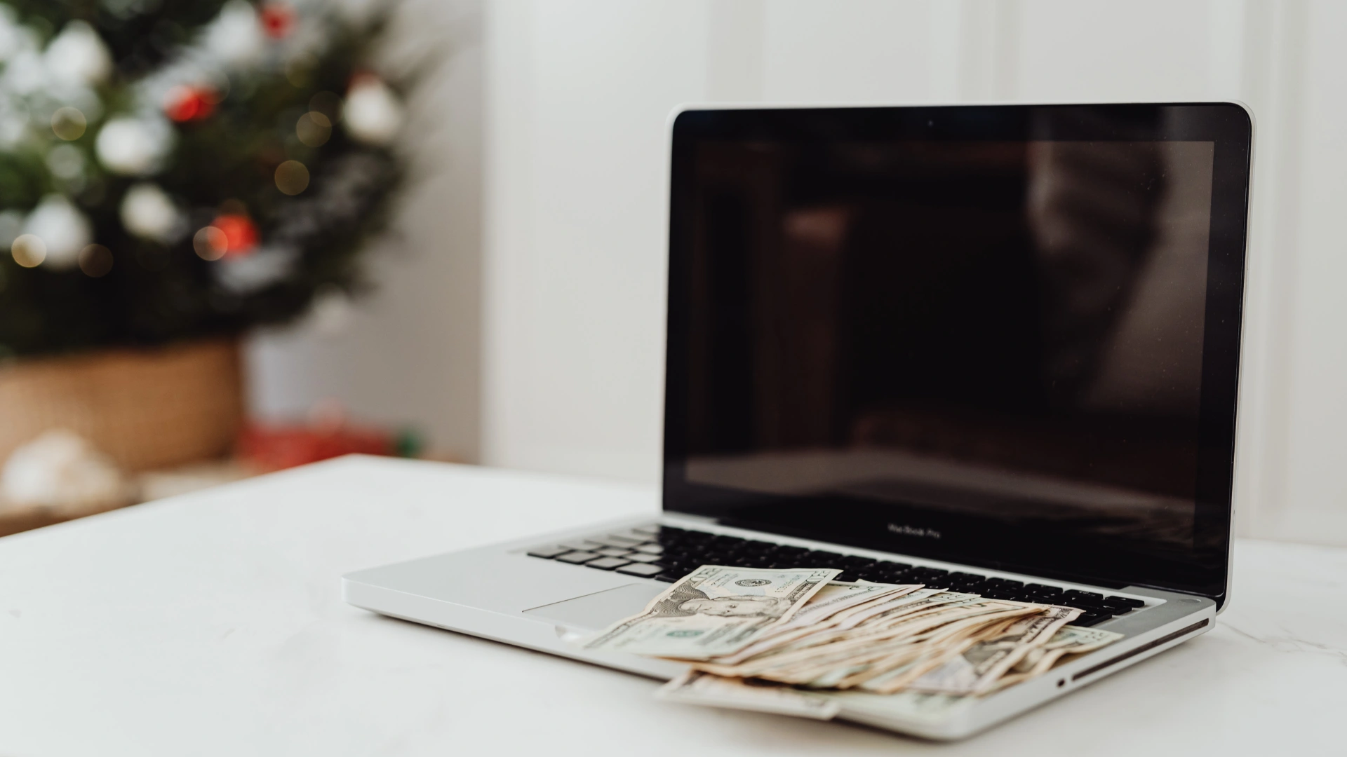 A laptop with U.S. dollar bills on its keyboard sits in front of a blurred Christmas tree.
