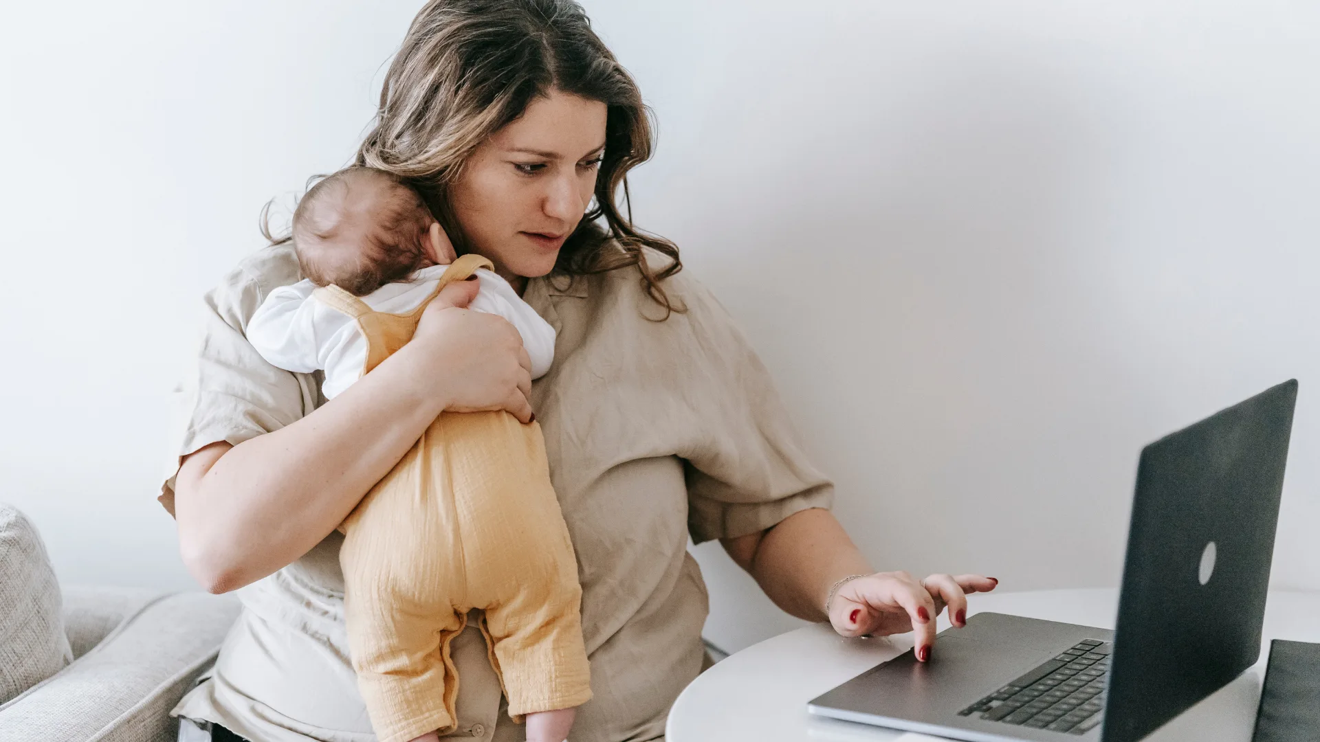 Mother holding baby while working on a laptop.