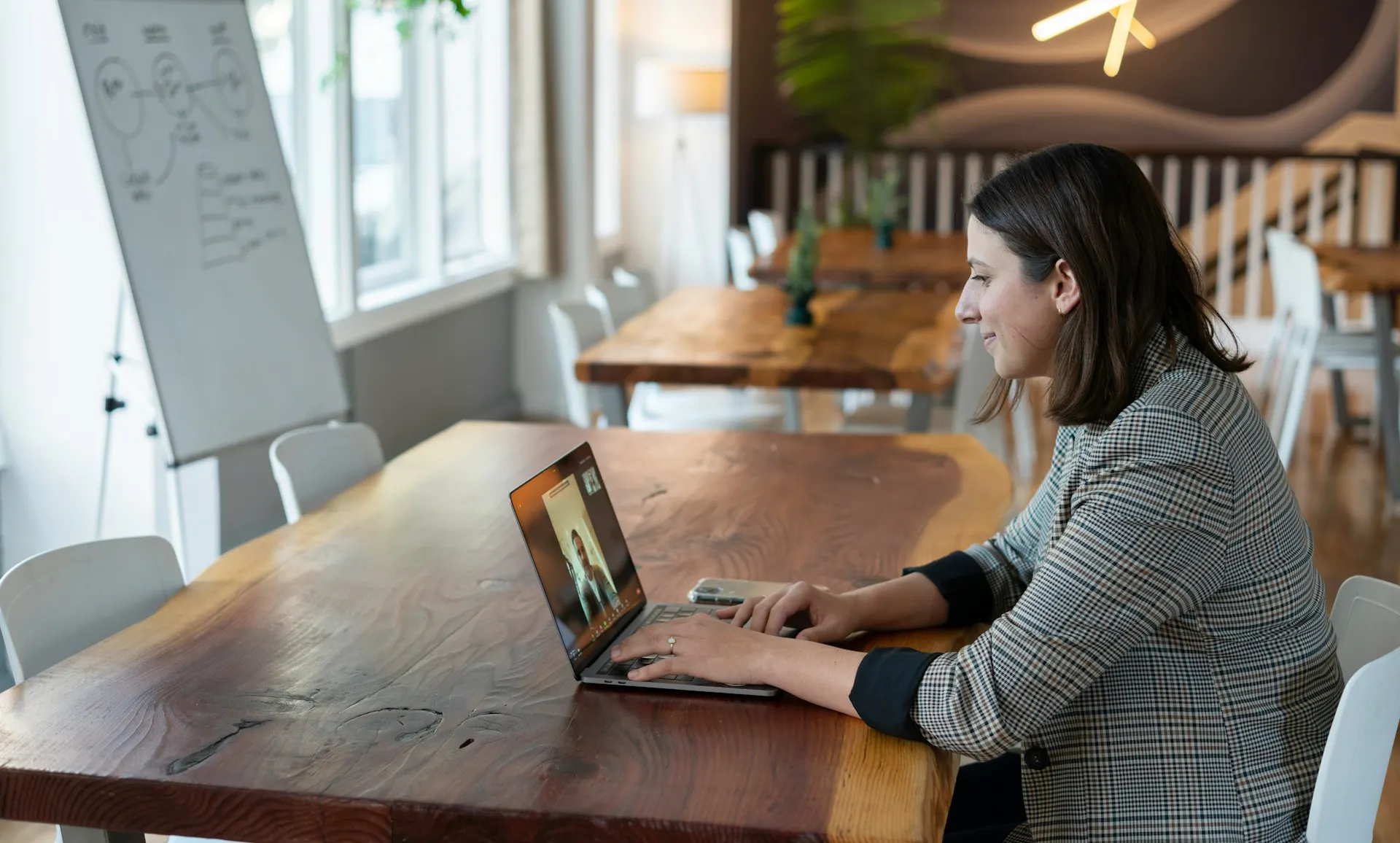 A woman in a plaid jacket video calls on her laptop at a large wooden table in a modern office space.