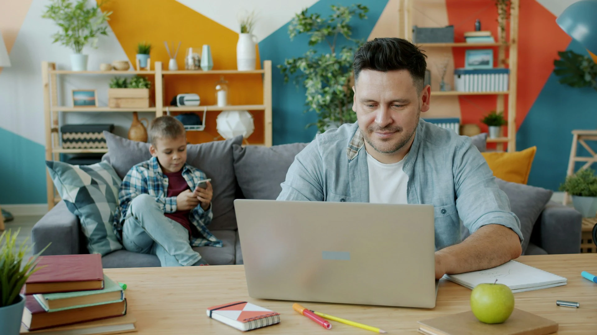 Father works on a laptop while son uses a smartphone on the couch in a colorful living room.
