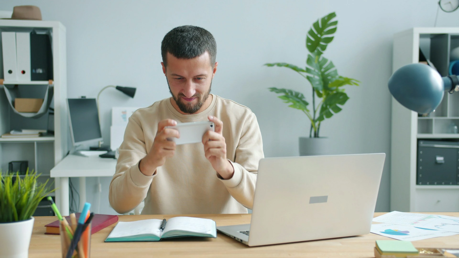 Man in a tan sweatshirt holds a smartphone horizontally in front of a laptop on a desk.