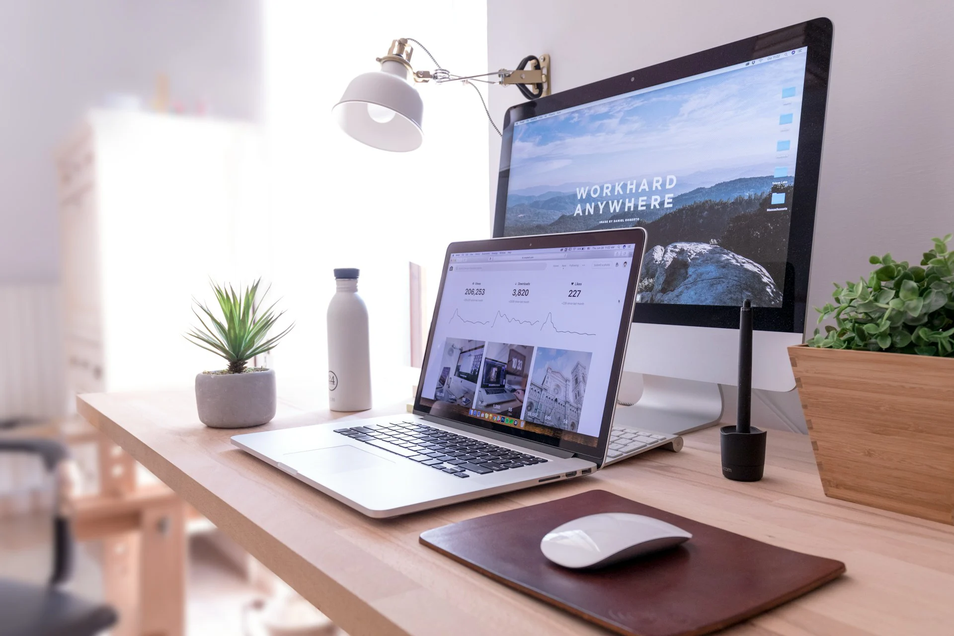 A modern home office desk setup featuring an open laptop, desktop monitor displaying "WORKHARD ANYWHERE", a water bottle, and potted plants.