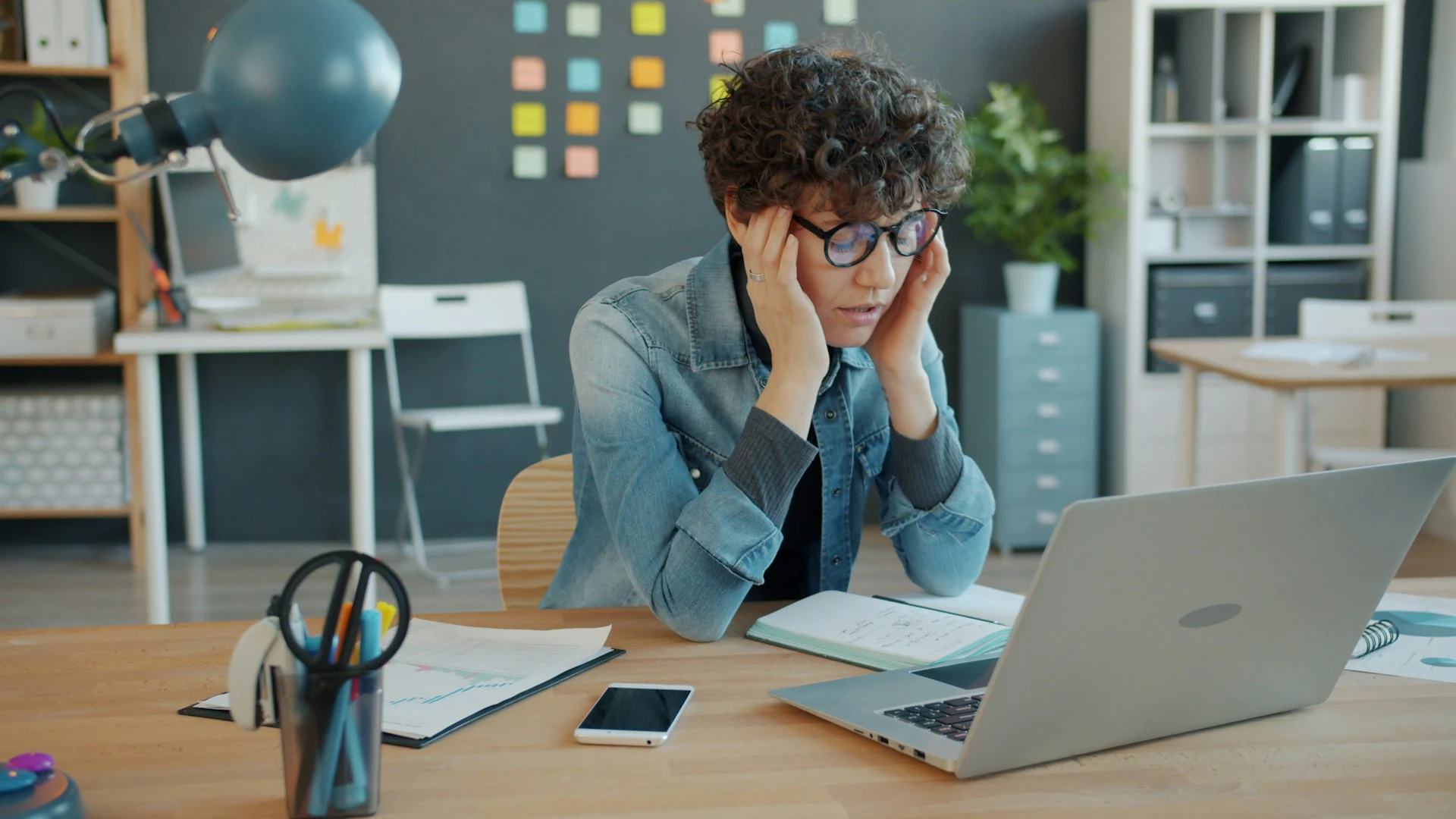 A stressed woman with curly hair and glasses sits at a desk with a laptop and papers, holding her head in her hands.