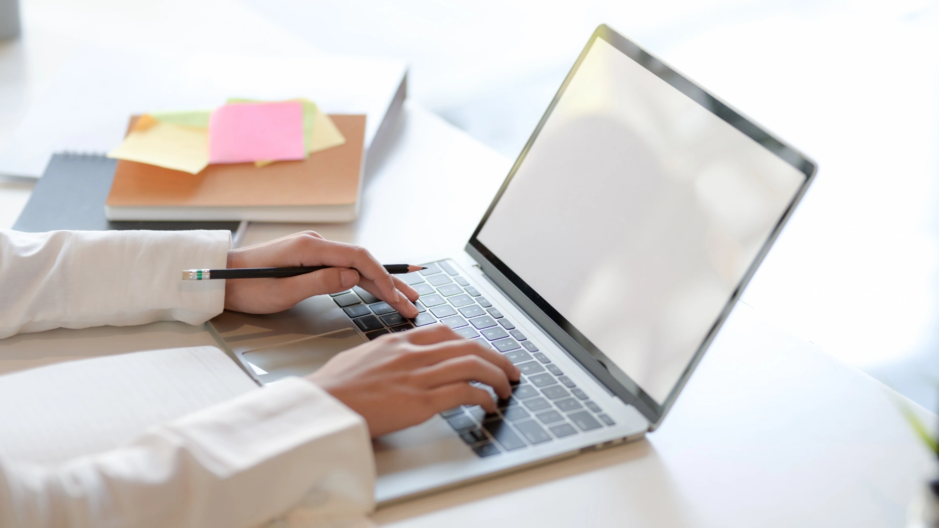 Hands typing on a laptop keyboard next to a notebook and colorful sticky notes.