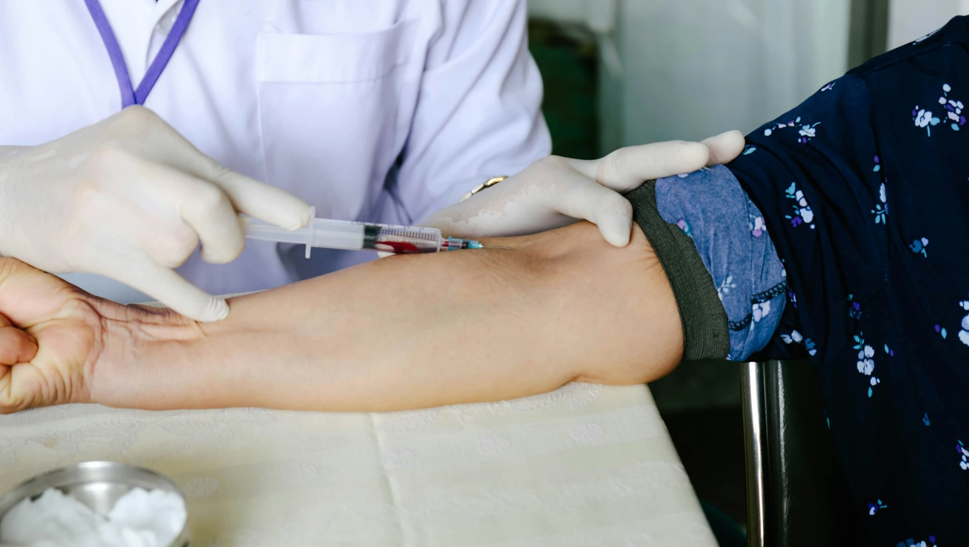 A medical professional in gloves draws blood from a patient's arm with a syringe.