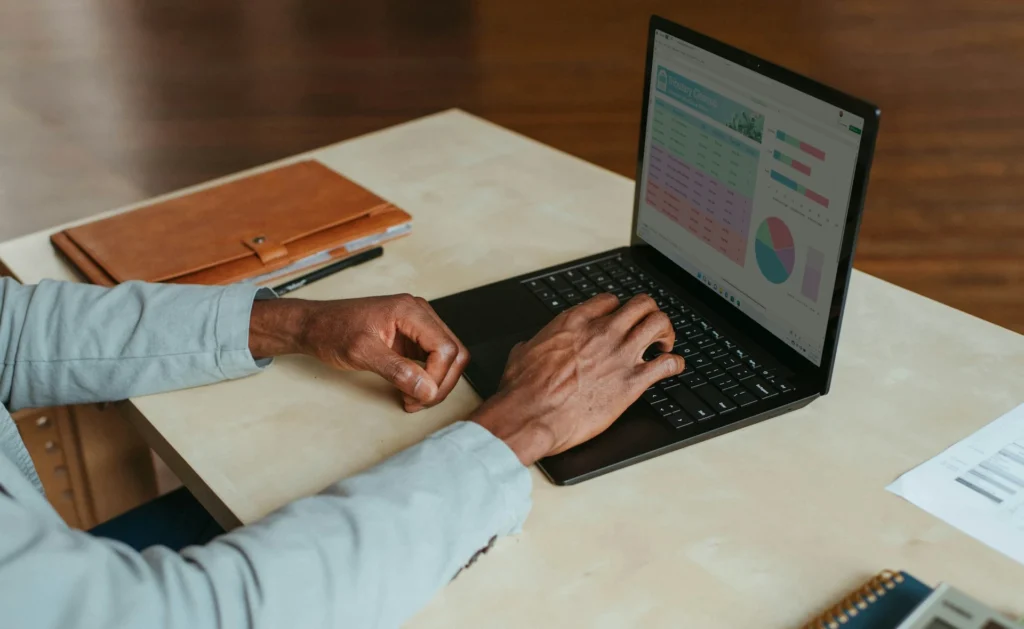 Man's hands typing on a laptop displaying financial charts and graphs next to a brown leather notebook.