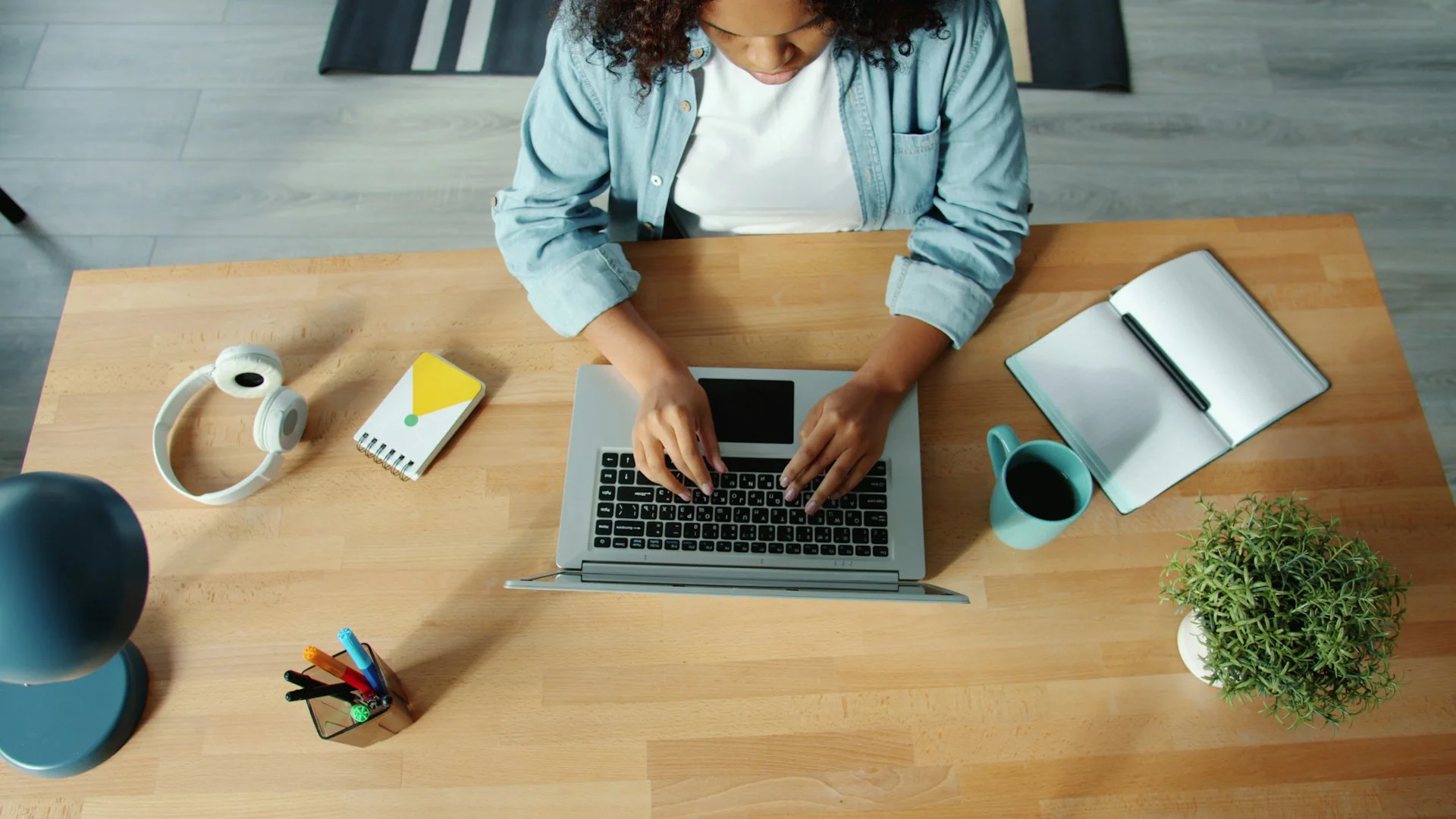 Overhead view of a woman typing on a laptop at a wooden desk with headphones, notebook, mug, and plant.