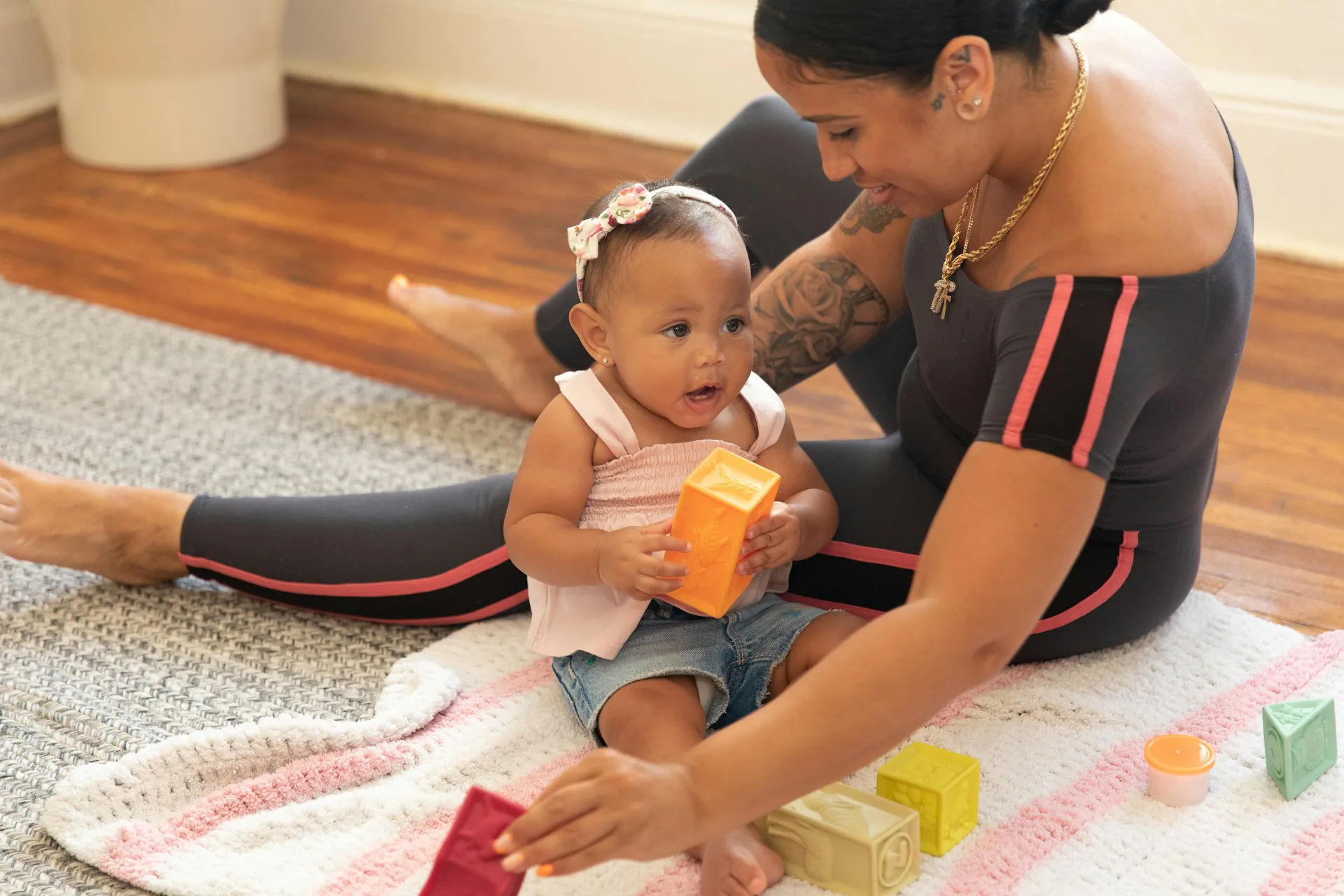 Tattooed mother playing with her baby on a soft rug as the child holds an orange cup among colorful toy blocks.